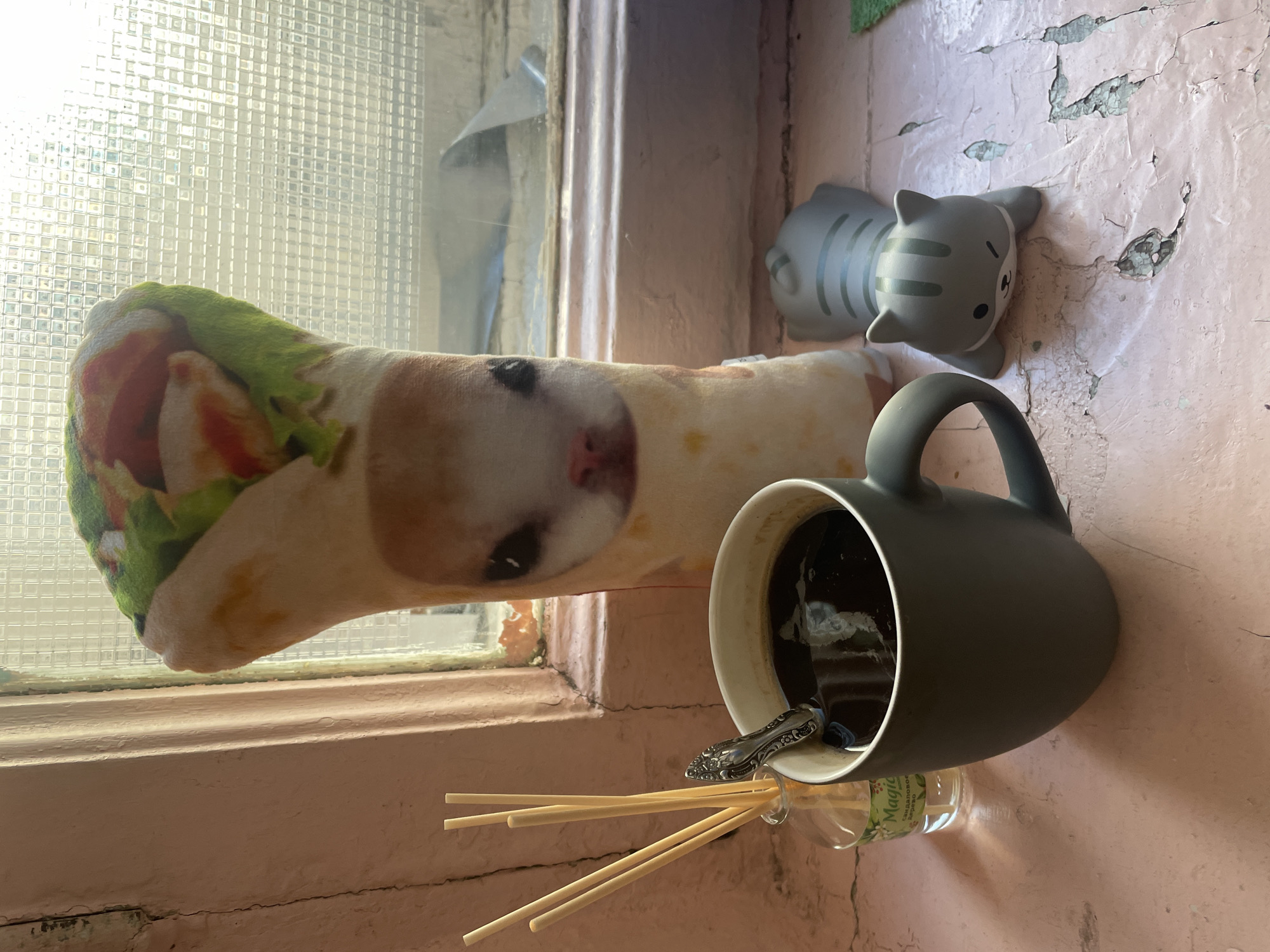 A close-up, eye-level shot captures a still life arrangement on a weathered, pale pink windowsill, likely in Saint Petersburg, Russia, given the style of the window and the likely location of the "Magic" brand diffuser. In the foreground, a dark grey mug filled with black coffee sits to the left, a silver spoon resting on its rim. To the left of the mug, a small glass bottle of "Magic" brand diffuser oil with a green label and several thin wooden reeds are positioned upright. Behind the mug, a plush toy resembling a burrito with a cat's face printed on it stands vertically. To the right of the mug and slightly behind it, a small, grey, stylized cat figurine with dark stripes and a wink lies on its belly. The background consists of a grimy, textured window with a grid pattern, through which diffused daylight streams, illuminating the scene. The windowsill itself is visibly cracked and peeling, indicating age and exposure.