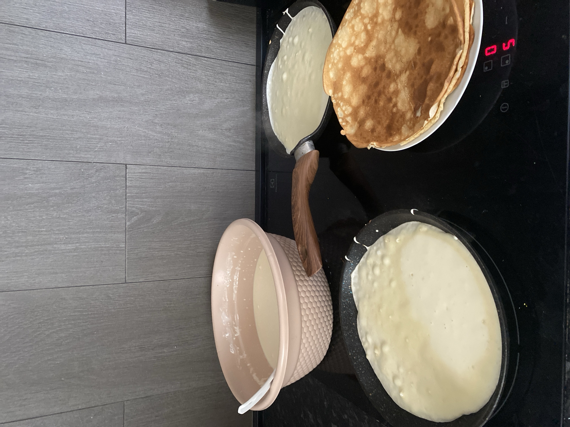 The image shows a kitchen counter with a black stovetop. On the stovetop, there are three pans. Two pans contain pancake batter, one is being cooked and the other is ready to be flipped. A stack of cooked pancakes sits on a plate next to the pans. A large bowl of pancake batter is also visible on the counter. The background features a gray tiled wall. The scene appears to be in a domestic kitchen.