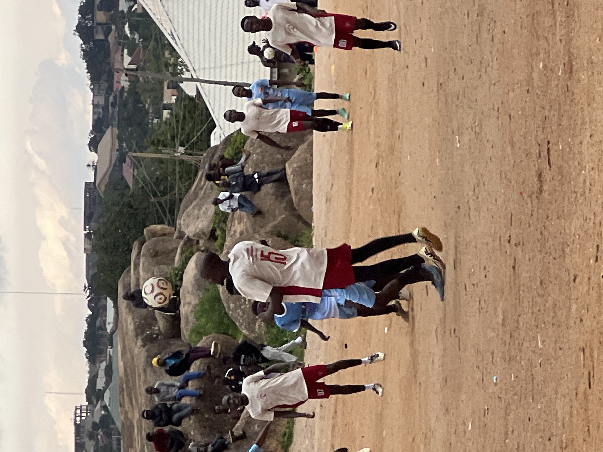 The image depicts a group of young men playing soccer on a dirt field in Jos, Nigeria. The scene is outdoors, with buildings and trees visible in the background, suggesting an urban or semi-urban environment. The sky is overcast, indicating a potentially cloudy or impending rainy day.
The primary subjects are the male soccer players, dressed in athletic attire, mostly white jerseys with red shorts or vice versa. Some players are actively engaged in the game, with one player in the foreground appearing to kick a soccer ball. Another player in the mid-ground is also in motion. Spectators are scattered around the field, some sitting on large rocks that are a prominent feature of the landscape. These rocks add a unique geographical element to the location.
The overall mood seems casual and recreational, with the players focused on their game and the spectators observing. The time of day appears to be late afternoon or early evening, given the lighting conditions.
Visible text includes the number "10" on the back of a white jersey worn by one of the players. The overall impression is of a community enjoying a sporting activity amidst a distinct local landscape.
