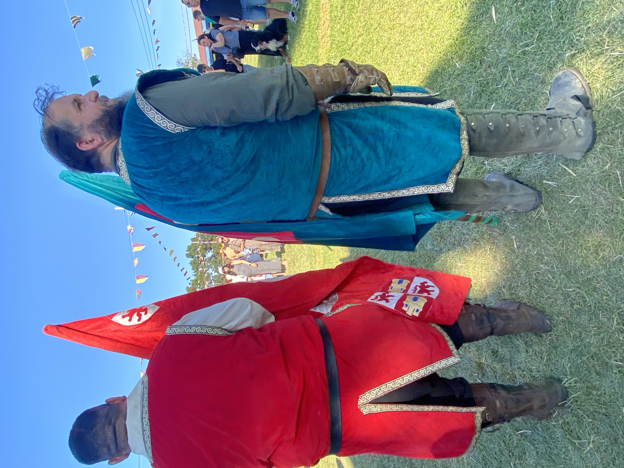 Two men in medieval costumes stand on a grassy field under a clear blue sky. The man on the left wears a bright red tunic with ornate trim and dark trousers, holding a red flag with a white emblem. He wears dark leather boots. The man on the right is dressed in a teal tunic with decorative trim over a green undershirt, and wears a leather belt and gauntlets. He stands with his back to the camera, facing right, and wears tall, grey leather boots. A teal flag is partially visible behind him. In the background, a crowd of people are gathered, some standing and others sitting, with a dog lying down on the grass. Colorful triangular flags are strung across the sky between poles. The scene appears to be an outdoor event, possibly a historical reenactment or festival, and the image location is identified as Llanera, Spain.