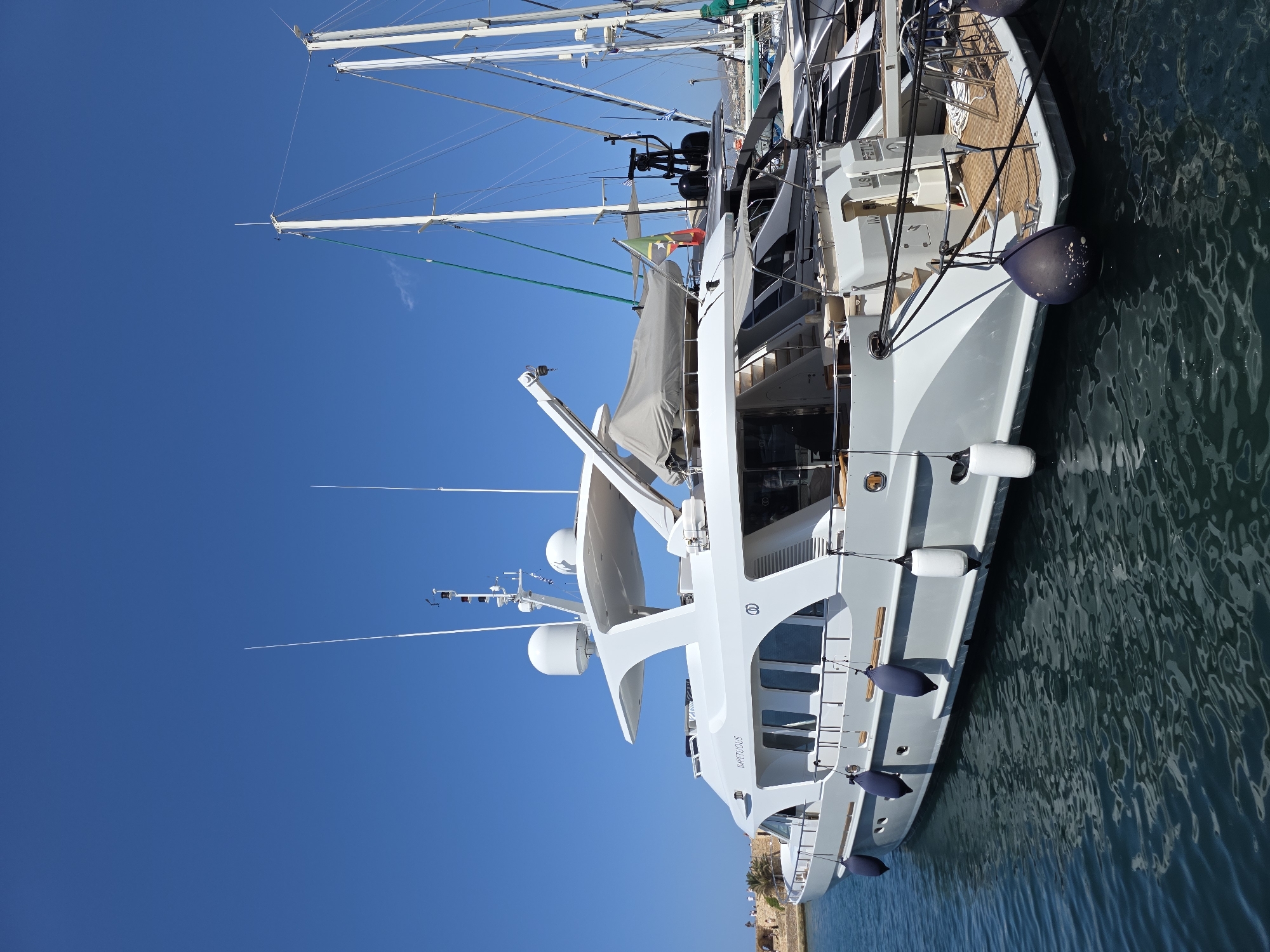 A wide, low-angle, eye-level shot captures a white motor yacht moored in the harbor of Chania, Greece. The yacht, named "IMPETUOUS," is positioned in the foreground, occupying the majority of the frame. Its sleek, modern design is emphasized by its pristine white hull and superstructure. Several dark blue fenders hang from its port side, cushioning it against the harbor wall. A white fender is also visible on the aft port side. The yacht's deck is made of warm-toned wood, and various ropes secure it to the dock.
Behind the "IMPETUOUS," several other boats, primarily sailboats with tall masts, are moored. The masts of these sailboats create a dense, vertical forest against the clear, vibrant blue sky, which takes up the upper portion of the image. A few small clouds are barely visible in the far distance.
To the left of the yacht, in the background, part of Chania's old Venetian harbor wall is visible, constructed of rough, weathered stone. A few palm trees are also present along the shoreline, suggesting a Mediterranean climate. A small group of people can be seen walking along the top of the harbor wall, providing a sense of scale. The water in the harbor is a deep, dark blue-green, with subtle ripples indicating a calm day. The overall scene depicts a sunny, peaceful day at a maritime location.