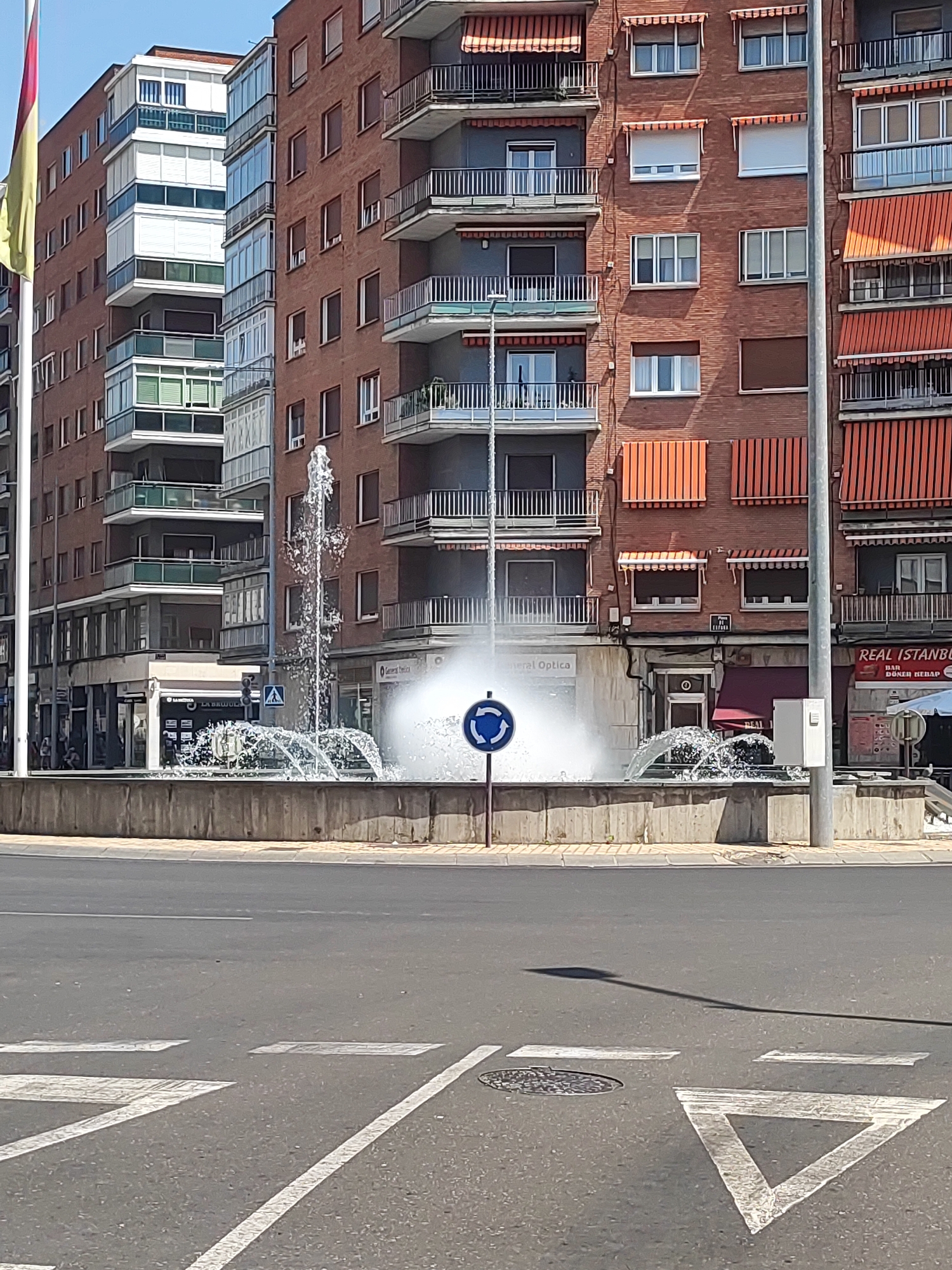 The image depicts a street-level view of a roundabout in Palencia, Spain, featuring a public fountain with water jets. The fountain is situated within a concrete basin. Several modern multi-story buildings with balconies and awnings line the background. One building displays a sign for "REAL ISTANBUL BAR DONER KEBAB". Traffic markings, including white lines and a triangular yield sign painted on the asphalt, are visible in the foreground. A blue circular traffic sign with arrows indicating a roundabout is placed near the fountain. The sky is clear and blue. No people are clearly visible or interacting with the scene.
