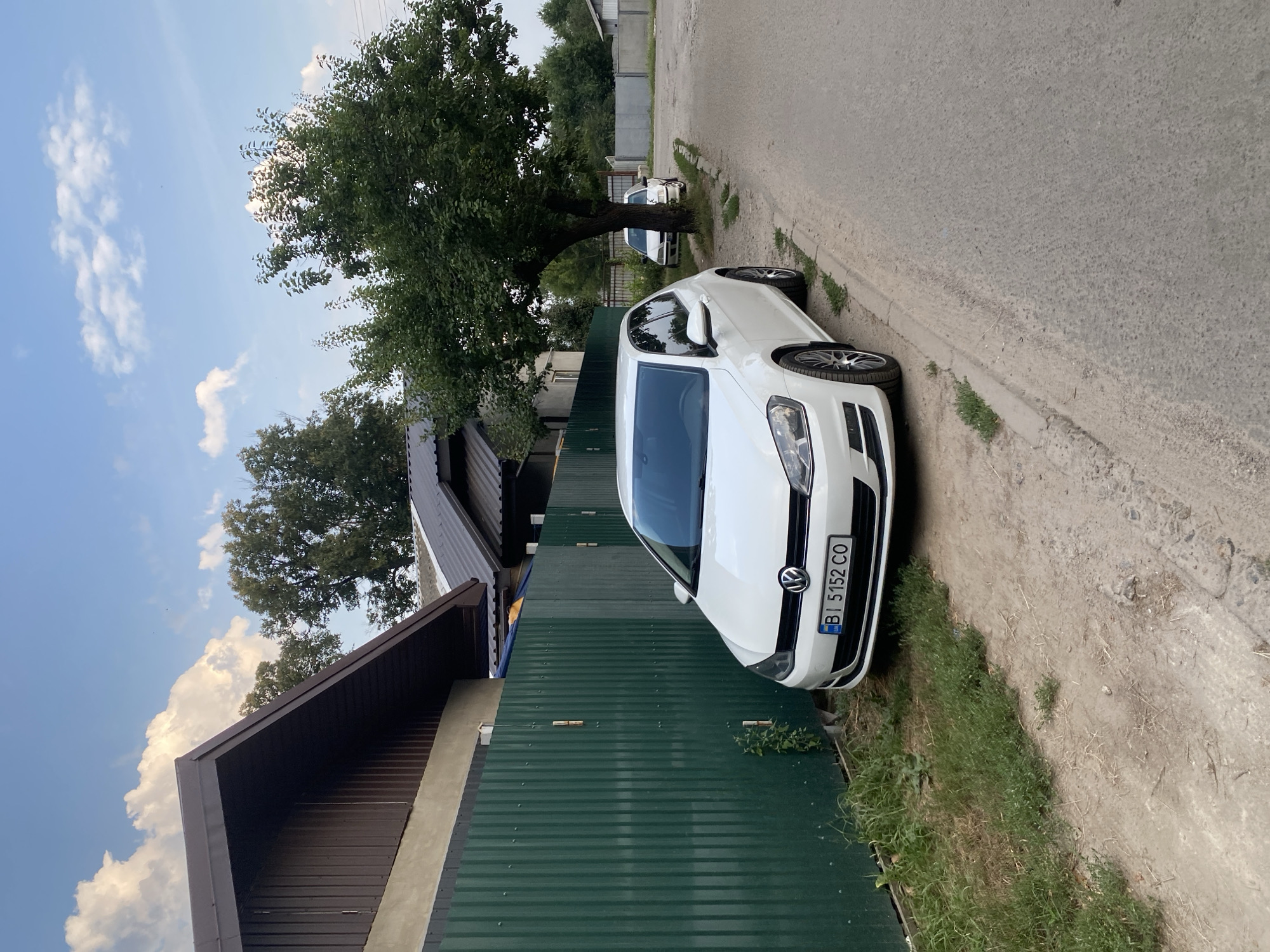 The image captures a white Volkswagen Golf parked on the side of a paved road in Poltava, Ukraine. The car is positioned close to a tall, dark green corrugated metal fence that runs alongside a property. Beyond the fence, a building with a dark brown metal roof is visible. A large tree with dense green foliage stands to the right of the car, casting shadows on the road and the car itself. In the background, another white car is parked further down the road, and more trees and buildings can be seen. The sky is partly cloudy with a mix of blue sky and white clouds. The overall environment appears to be a residential or semi-rural area. The license plate of the white Volkswagen Golf is clearly visible and reads "BI 5152 CO".