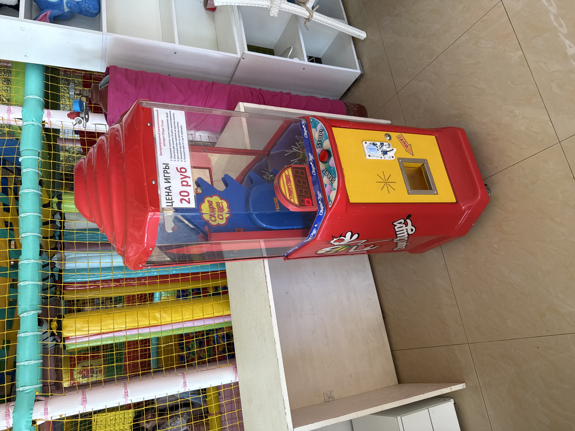 This is a medium shot, eye-level, interior photograph of a Chupa Chups dispensing machine in a children's play area. The machine is red and yellow and is positioned on a tiled floor. To the left of the machine is a white, wooden counter with rounded corners. Behind the machine and counter is a brightly colored play structure with yellow, pink, and blue padding and yellow netting. A large pink fabric roll is to the right of the machine, and white shelving units are further to the right. The Chupa Chups machine has a glass front revealing a mechanism with what appear to be prizes. A digital display shows "8888". Text on the machine reads "ЦЕНА ИГРЫ 20 руб" (Price of the game 20 rubles) and instructions in Russian. The word "START" is visible on a red button, and "STOP!" is on a yellow button. A small square opening with an arrow pointing to it is labeled "OUT!". The overall environment suggests a commercial establishment like a family entertainment center or a shopping mall play zone. The city of Sochi, Russia, is mentioned, which is consistent with the Cyrillic text and the style of the play area. There are no people clearly visible in the frame.