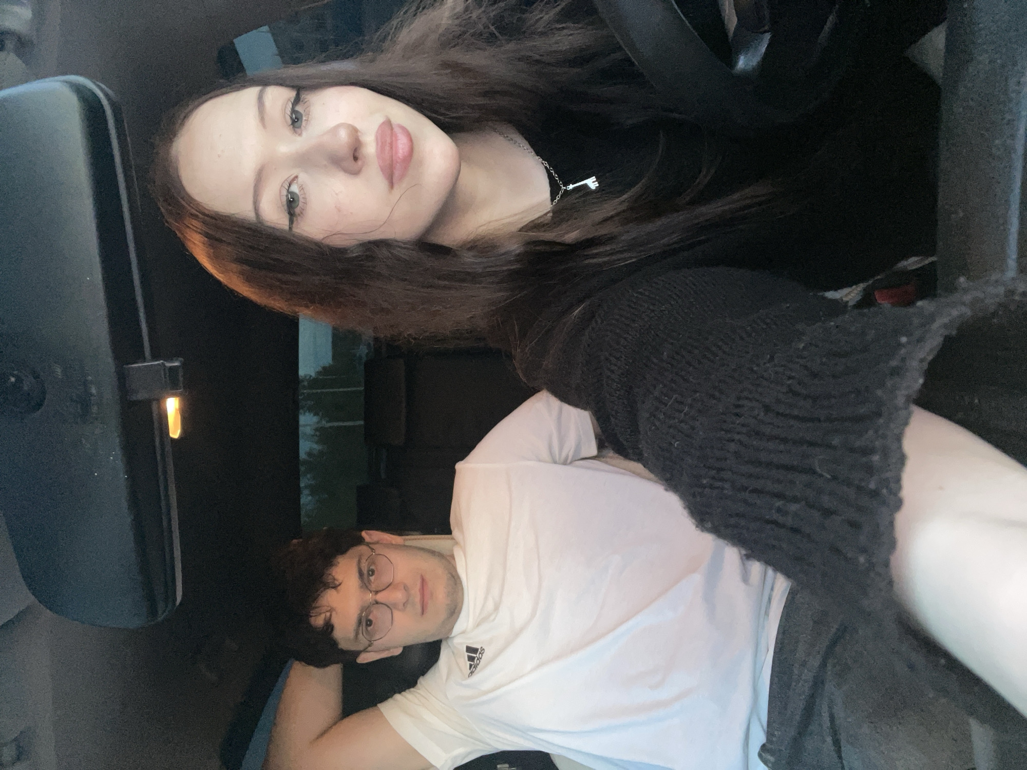 The image displays two individuals, a woman and a man, inside what appears to be a car, oriented vertically as if taken as a selfie or portrait.
The woman is positioned in the upper part of the frame, with long, dark brown hair flowing downwards. She looks directly at the camera with a neutral to slightly pouting expression. She has light-colored eyes accented with eyeliner and full lips. She is wearing a dark, possibly black, top and a silver necklace featuring a distinctive, elongated, stylized pendant. Her arm, covered by a dark, possibly knitted sleeve, is visible on the right side of the frame.
The man is situated below and to the right of the woman, appearing to be lying down or leaning back. He has short, dark, curly hair and is wearing round, thin-framed glasses. He looks towards the camera with a calm, neutral expression. He is dressed in a white t-shirt, where the "Adidas" logo is clearly visible on his left chest. One of his arms is bent, seemingly supporting his head or resting comfortably.
The setting is the interior of a vehicle. A rearview mirror is prominent on the left side of the frame, showing some reflection. The interior appears dark, suggesting low light conditions, possibly evening or a heavily shaded area. The background visible through the car windows is indistinct.
The overall impression is that of a casual photo or selfie taken by the individuals. Both subjects maintain composed expressions while engaging with the camera. The location context provided is Votkinsk, Russia.