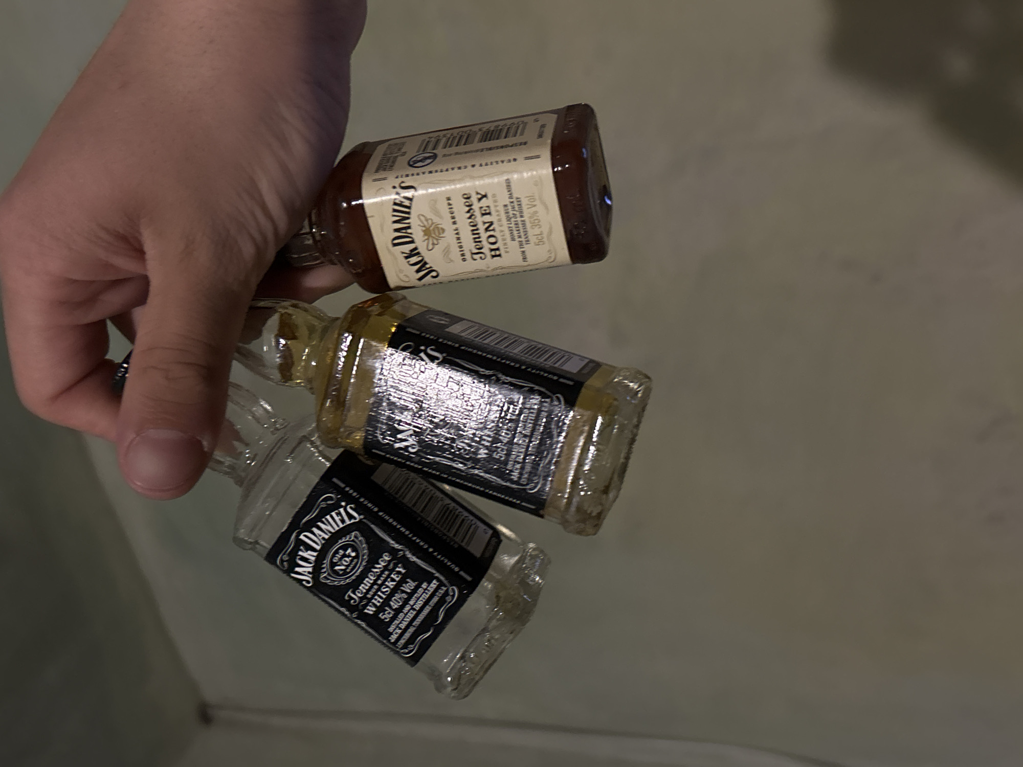 The image shows a hand holding three miniature bottles of Jack Daniel's whiskey. From top to bottom, the first bottle is Jack Daniel's Tennessee Honey, the second and third bottles are Jack Daniel's Old No.7 Tennessee Whiskey. The scene is indoors, possibly a residence. There is no indication of Yerevan, Armenia, in the image.