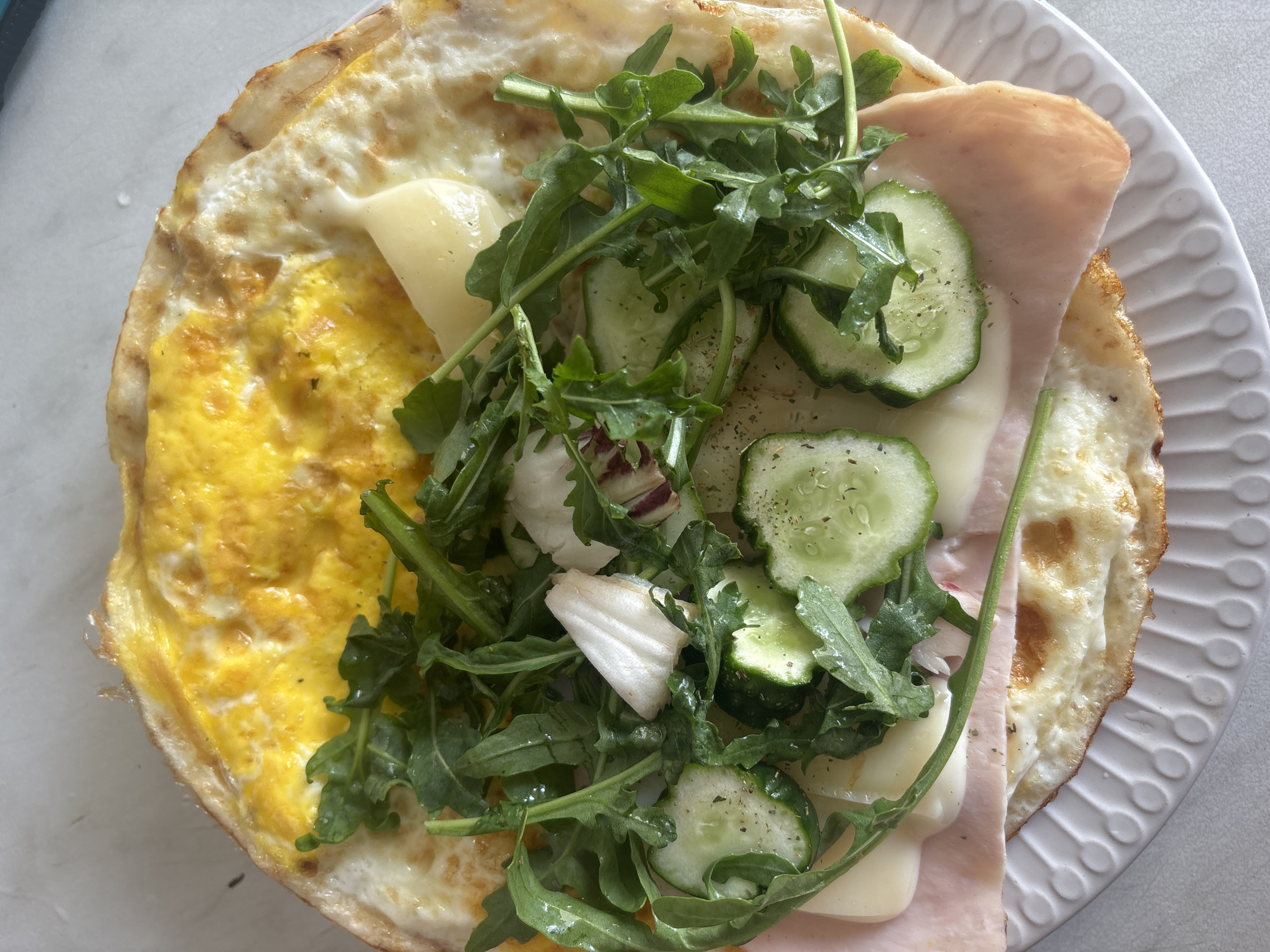 A close-up, top-down shot showcases a disassembled sandwich or wrap presented on a white, textured plate. The primary components include a fried egg, thinly sliced ham, melted cheese, and fresh greens, specifically arugula and some red leaf lettuce, interspersed with sliced cucumbers. A dollop of a light-colored sauce, possibly mayonnaise or aioli, is visible atop the greens. The overall presentation suggests a breakfast or light meal. The scene is devoid of any context relating to the city of Klin, Russia, or any identifiable environmental elements that could place it there.