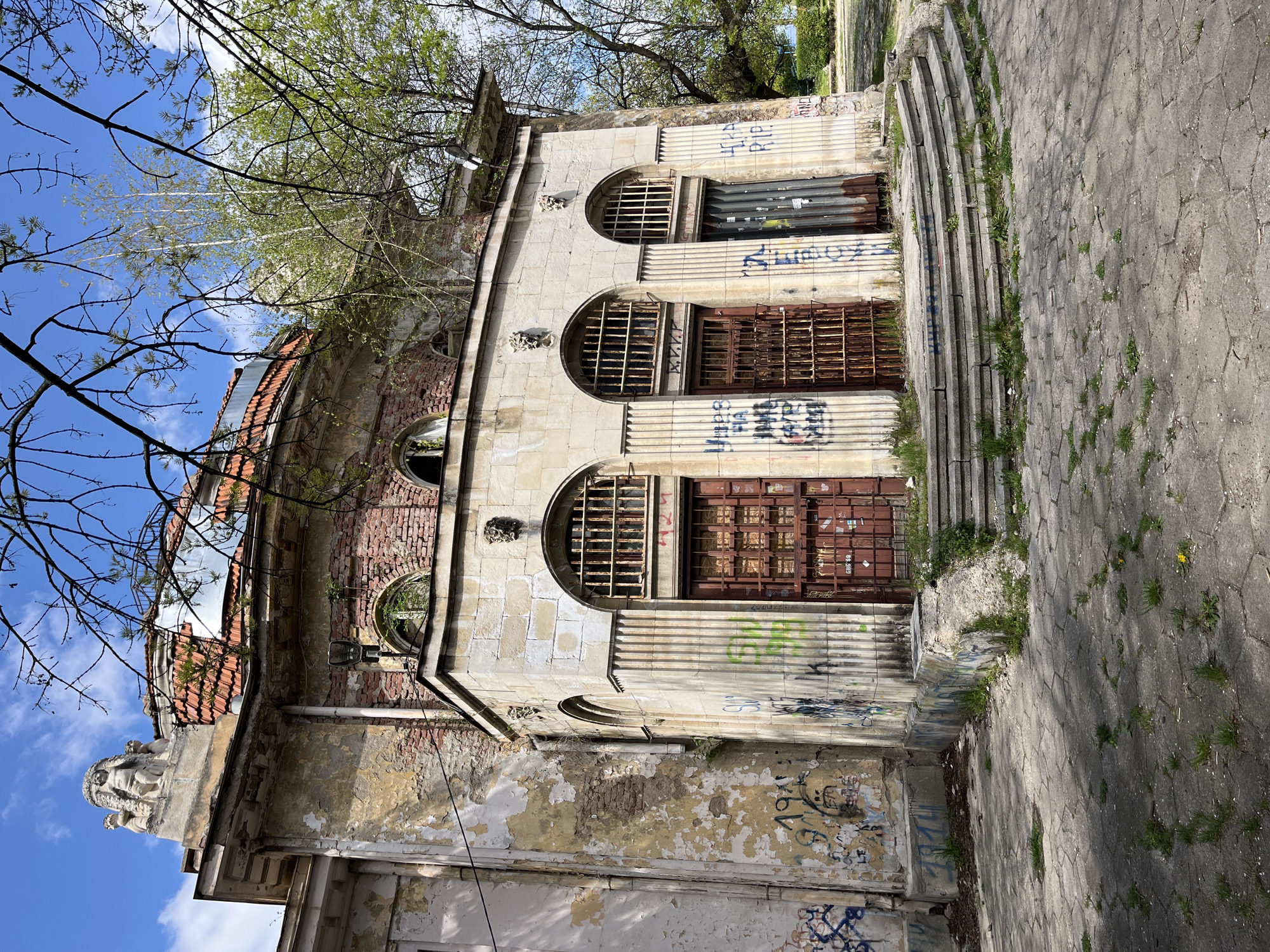 This photograph captures an old, dilapidated building viewed from a low angle, giving it a grand, albeit decaying, presence. The building features classical architectural elements, including prominent fluted pilasters and arched window openings. The facade is constructed from stone blocks, with sections revealing exposed red brickwork and peeling, faded paint.
The structure is in a state of neglect, with visible signs of deterioration. Several arched windows are covered by rusty metal grates, while others have corrugated metal doors or shutters, also rusted and covered in graffiti. Small, decorative stone sculptures, resembling gargoyles or stylized heads, are placed above some of the arches. On the far left, near what would be the roofline if the image were upright, a larger, ornate sculpture of a bearded figure is visible.
The surrounding area includes a paved path of hexagonal concrete blocks, leading to a series of curved concrete steps that ascend alongside the building. Weeds and grass grow through cracks in the pavement and steps, further indicating disuse. Overhanging tree branches with bare twigs and new green leaves frame the bright blue sky, which has a few scattered white clouds. The clear light suggests a sunny daytime setting.
Numerous graffiti tags and words are scrawled across the building's facade, particularly on the metal covers of the windows and the lower walls. Recognizable text includes "RIP", "HIRA", "PIRLO", and "V.V.V.", among other illegible markings. There are no people present, and no specific activity or event is taking place, contributing to the sense of abandonment and forgotten history.