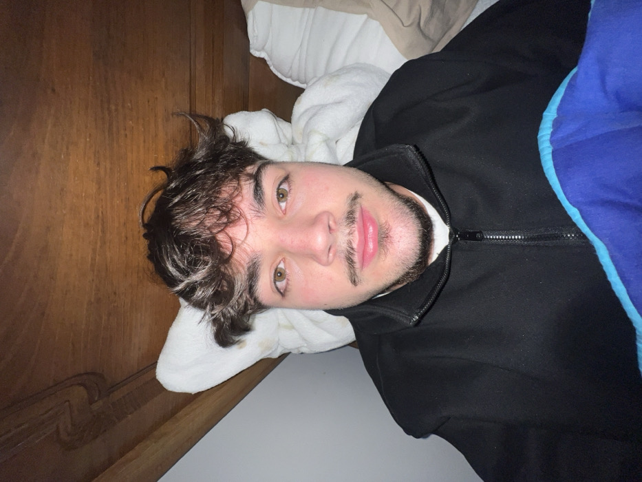 The image features a young adult male positioned in an indoor setting, likely a bed. The subject has dark, curly hair, light brown eyes, a light complexion, a moustache, and a goatee. He is wearing a black zip-up jacket over a white garment, with a blue blanket or duvet featuring a light blue stripe covering the lower portion of his body. He is looking directly into the camera. Behind his head, a white pillow is partially visible, supported by a wooden structure, possibly a headboard or wall panel, which displays a carved detail on its upper left side. A plain white wall is visible to the left of the subject. The specific location within the city of Bragança, Portugal, cannot be determined from the visual information provided.