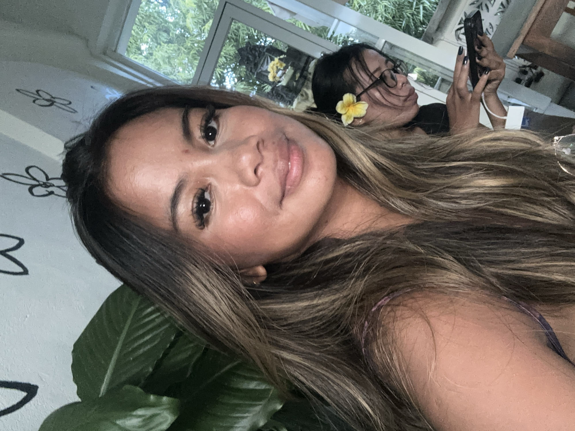 The image is a tilted selfie featuring two women in a relaxed indoor or semi-open setting, likely a cafe or restaurant in Ubud District, Indonesia. The primary subject, a woman in the foreground, looks towards the camera with a soft smile. She has long, dark brown hair with lighter highlights, styled to cascade over her shoulder, and is wearing visible makeup. A purple strap, possibly from her attire, is glimpsed on her shoulder.
Behind her, a second woman is partially visible, wearing glasses and a dark top. She has a bright yellow frangipani flower tucked behind her ear and is intently looking at her smartphone, which she holds with both hands. Her fingernails are painted a dark color.
The background shows large windows through which lush green foliage is visible, indicating a tropical or natural setting. The white walls or ceiling feature delicate black outline drawings of flowers. A large green plant leaf is prominent in the lower left foreground, adding to the natural aesthetic. The lighting suggests it is daytime, with ample natural light entering the space. The overall mood is casual and serene. No visible text is present in the image.