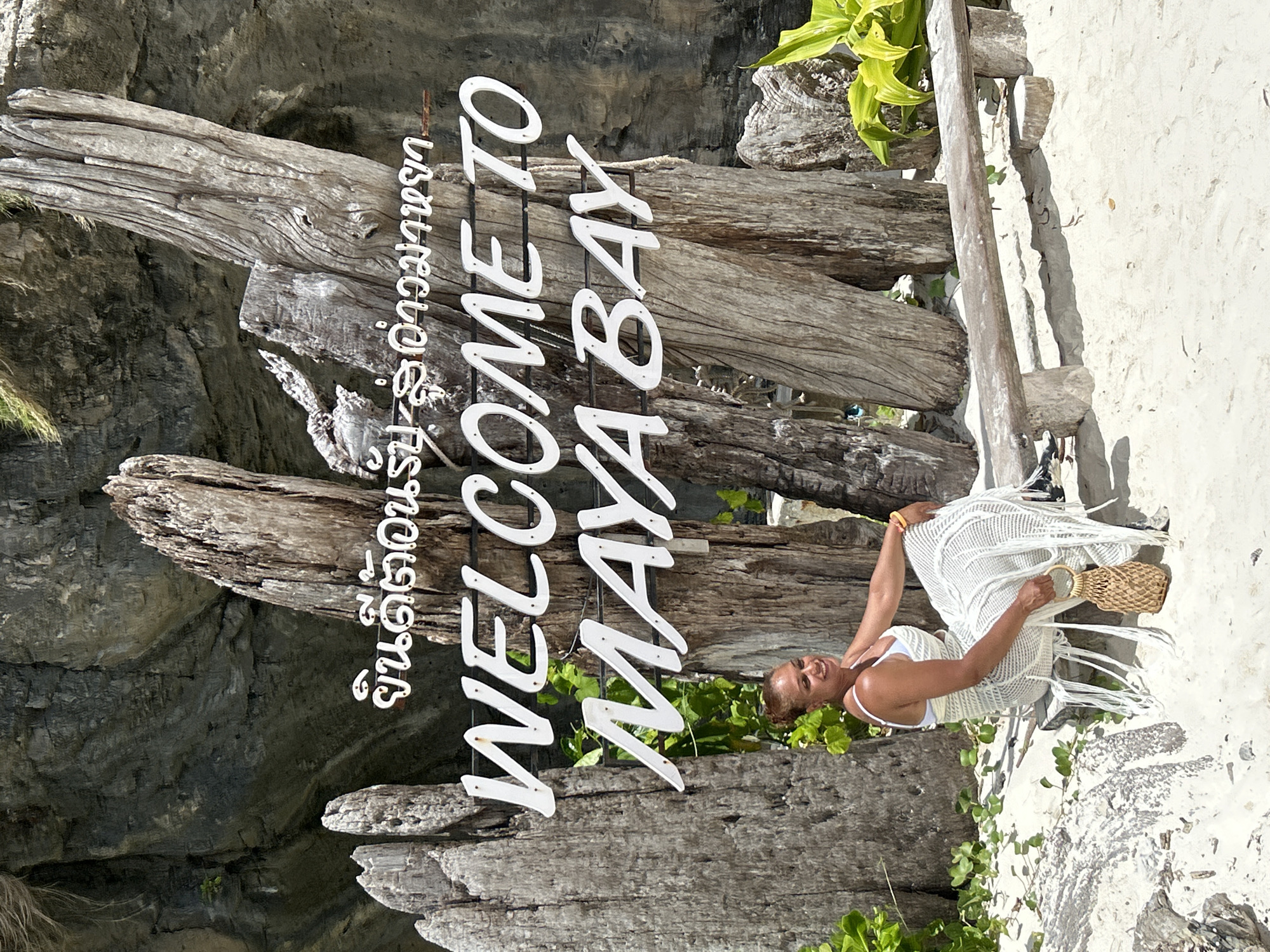 A full shot captures a woman sitting on a rustic wooden bench on a sandy beach, with a large, weathered sign that reads "WELCOME TO MAYA BAY" in large white letters. The sign is partially obscured by thick, gnarled pieces of driftwood, which are positioned vertically behind it. The background features a steep, gray rocky cliff face, with patches of green vegetation clinging to its surface and at its base. Lush green foliage also grows to the right of the sign, adding a vibrant contrast to the sandy environment.
The woman, who appears to be of mixed ethnicity, is wearing a white bikini top and a white crocheted dress with intricate fringe detailing. She is holding a small, woven straw bag with a circular wooden handle in her right hand. Her hair is pulled back in a bun, and she is smiling and looking to her left. She is seated on the left side of the bench, with her legs crossed.
The bench itself is made of rough-hewn wood and appears weathered by the elements. It is placed on the sand, with visible footprints around it. The sand is light in color and appears to be a fine grain. The overall scene is bathed in bright, natural sunlight, creating strong shadows. The environment is consistent with a tropical beach setting, and the "Maya Bay" sign specifically indicates the location is Maya Bay, which is near Ao Nang, Thailand.