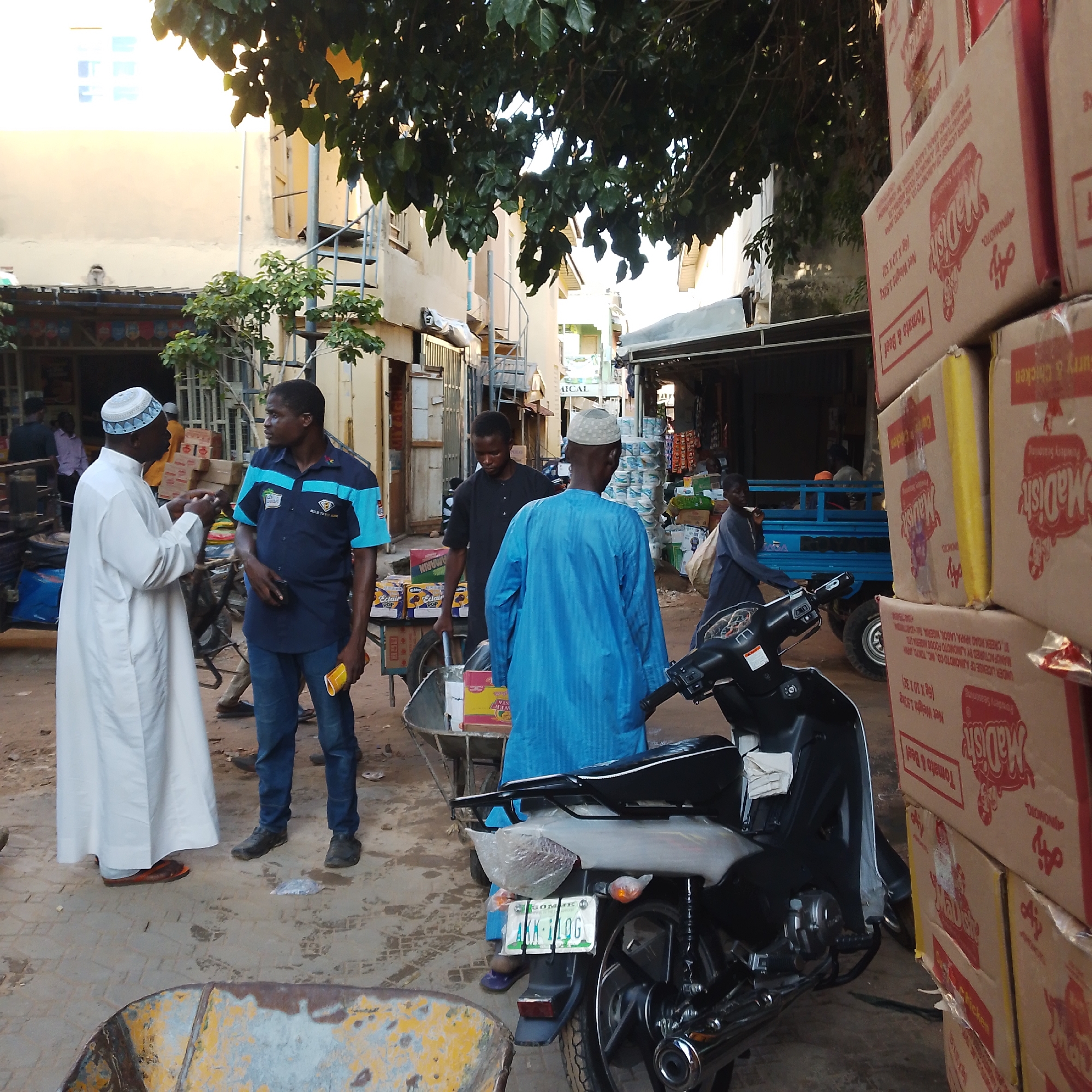 The image depicts a street scene in Arawa, Nigeria. In the foreground, a man in a white traditional outfit and a blue and white patterned cap is conversing with another man wearing a dark blue shirt with light blue accents and blue jeans. A motorcycle is parked in the lower right corner of the frame, with its license plate visible. Behind them, a wheelbarrow is filled with boxes. To the left, stacked boxes, some branded "Madish" with red text and illustrations, are prominently displayed, indicating a commercial area or market. In the background, there are buildings with an outdoor staircase, and several other people are visible in the distance, some appearing to be shopkeepers or customers. The environment suggests a bustling outdoor marketplace or street with shops lining the area. The lighting indicates it is daytime, with sunlight filtering through overhead foliage.