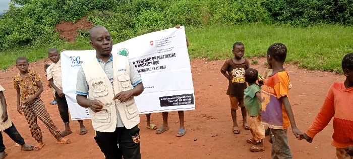 The image depicts an outdoor scene in Ngalula, Democratic Republic of the Congo, featuring one adult male and several children on reddish-brown dirt ground. The background consists of dense green vegetation, including trees and tall grass, with a reddish-brown soil mound visible on the left.
The adult male, centrally positioned, has dark skin and short, dark hair. He wears a grey t-shirt, dark trousers, and a beige multi-pocketed utility vest with text including "TPO/DRC" and "HUTTEK" partially visible. He stands facing slightly left, gesturing with his left hand and holding a small, indistinct object in his right hand, appearing to be addressing the group.
Behind the adult, two children hold a large white banner horizontally. The banner displays multiple logos, including one resembling the Swiss flag, and text in French and possibly Lingala, which includes "TPO/DRC UN PARTENAIRE," "S BAILLEUR RDC," and "AZEMBELEO YA NYAMI." An email address and two phone numbers are printed at the bottom.
At least seven children, ranging in age from young toddlers to pre-teens, are present. They wear varied casual clothing, some showing signs of wear. They are standing around the adult and banner, observing or looking in different directions. One child in the foreground on the right holds hands with another child. The scene is illuminated by natural daylight.