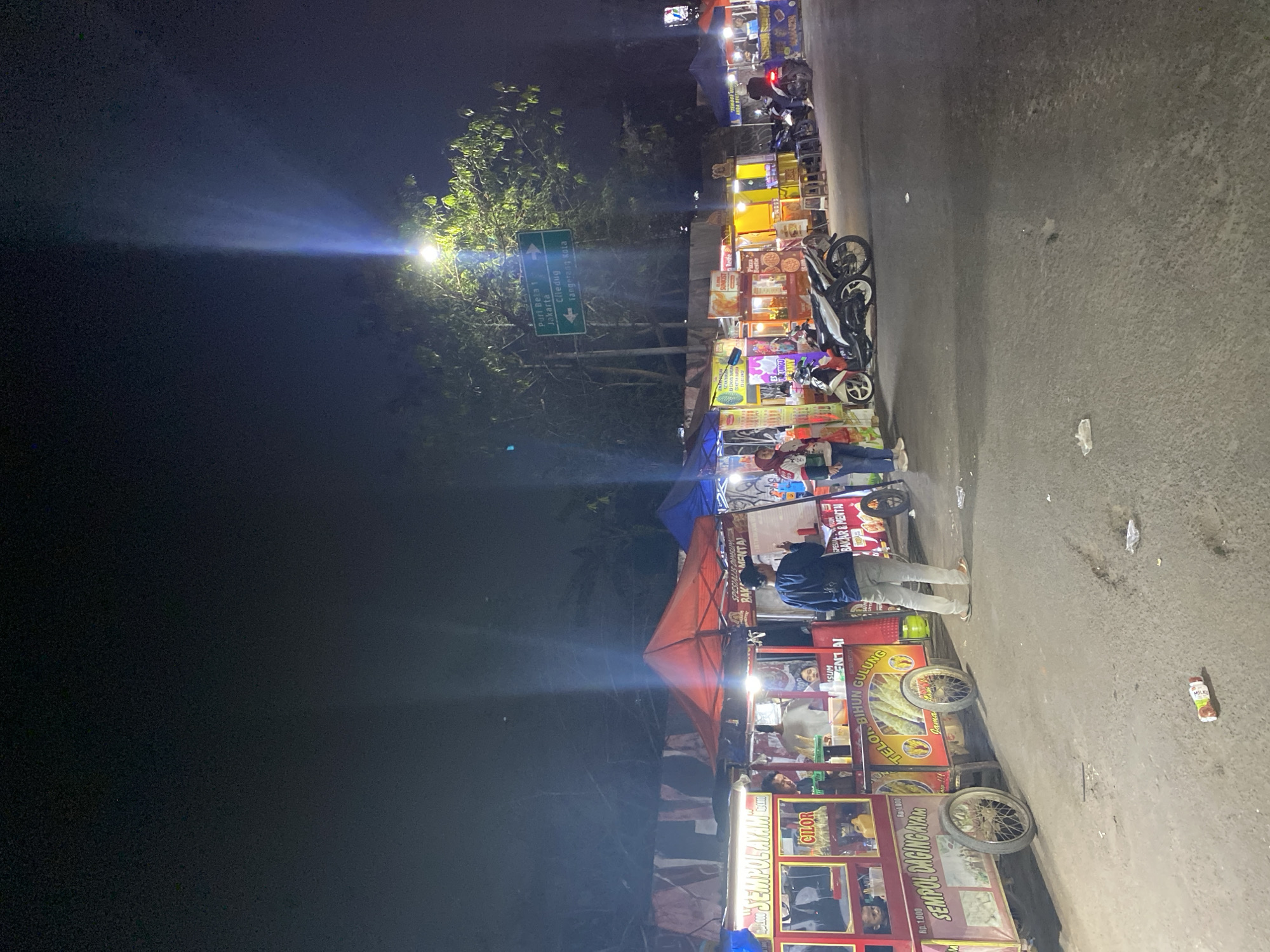 The image depicts a nighttime scene at a street food market, likely located in Tangerang, Indonesia, as suggested by a street sign indicating "Tangerang Kota." The market features several food stalls, some with bright lights illuminating their offerings. Various food carts, such as one labeled "SEMPOL AYAM" (Chicken Sate Skewers) and another with "TELUR BIHUN GULUNG" (Egg Spring Rolls), are visible. People are seen interacting around the stalls, some appearing to be customers and vendors. Motorbikes are parked in front of some stalls, indicating common local transportation. The ground is paved, with some debris scattered across it, typical of a public outdoor space. In the background, trees and a street light with a directional sign are present. The overall atmosphere suggests a lively, albeit simple, evening market environment.