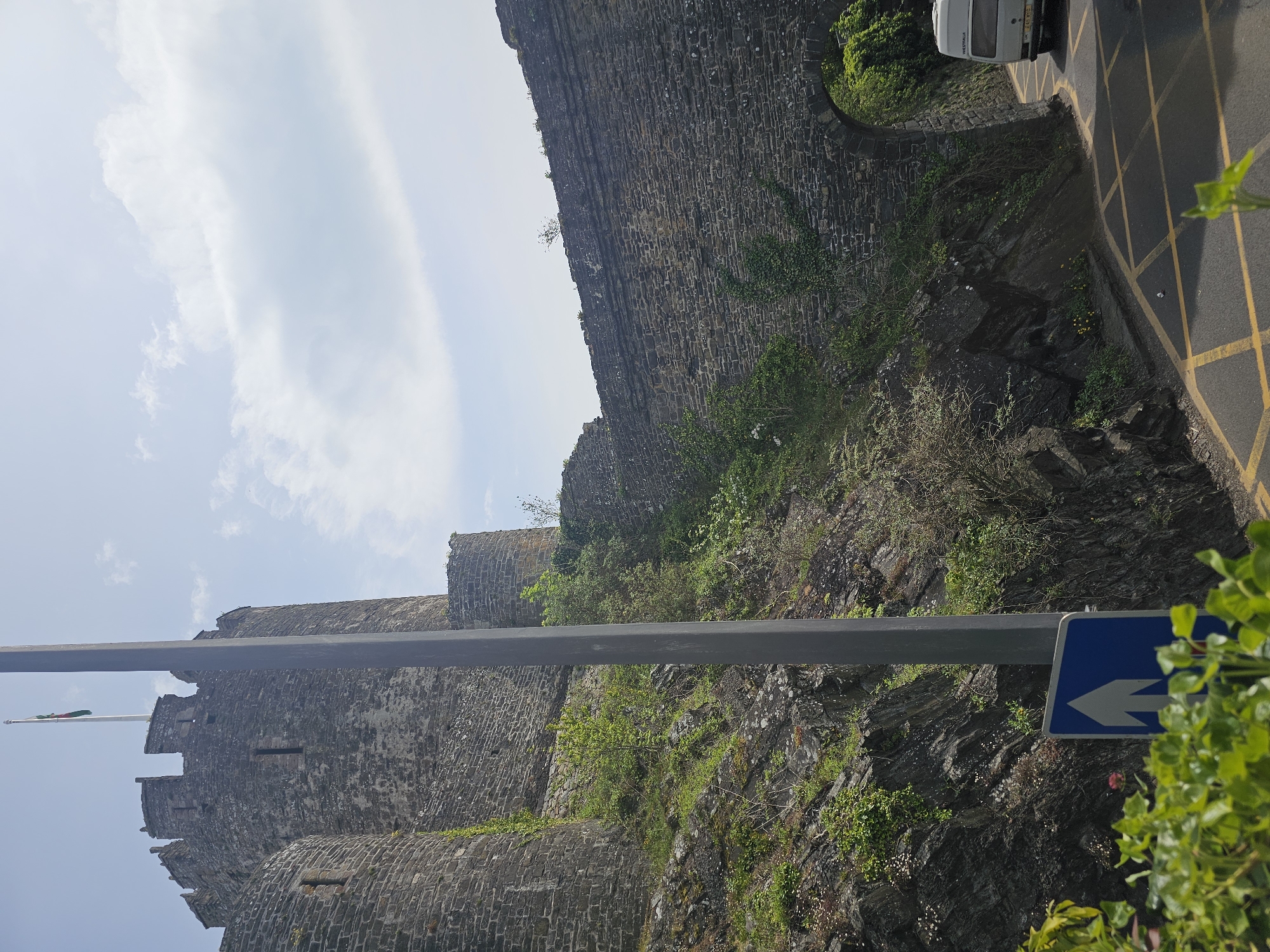 The image depicts a section of Conwy Castle in Llandudno Junction, United Kingdom, framed by a street sign and a roadside wall. The castle, built from stone, features crenellations and window openings. Vegetation grows on the castle walls and surrounding rock face. A road with double yellow lines and a parked white vehicle is visible at the top right. The sky is partly cloudy.