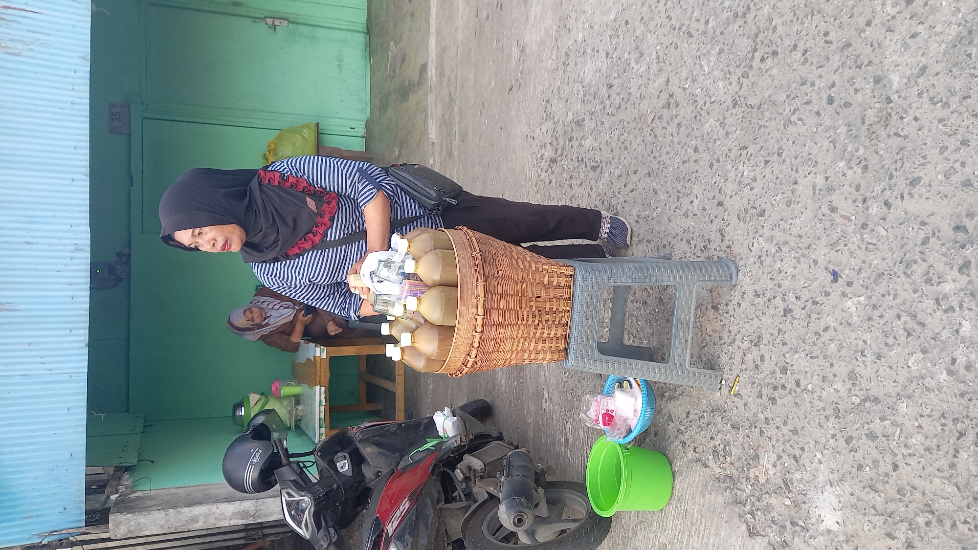 In Balikpapan, Indonesia, a woman wearing a black hijab and a striped shirt stands by a stand selling beverages from bottles in a woven basket. She is holding a bottle and a cup, and there are other bottles and cups in the basket. In the background, another woman in a hijab sits at a table. A motorcycle is parked to the left of the scene, and a green bucket and a blue basket are on the ground. The scene appears to be outdoors, possibly on a street or market.