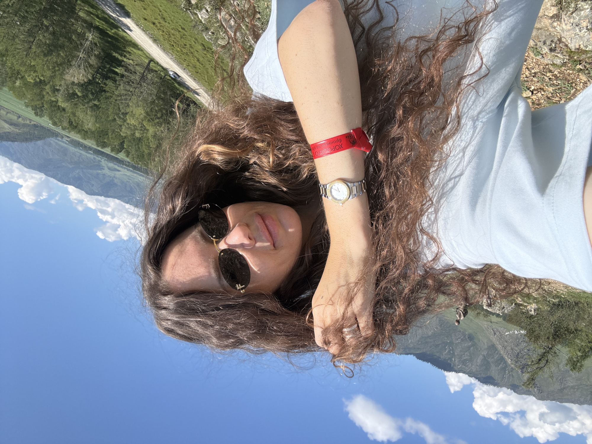 A selfie taken outdoors features a woman with long, curly brown hair and sunglasses. She is wearing a light blue t-shirt and a watch on her left wrist, along with a red wristband. Her hand is in her hair. The background shows a mountainous landscape with green slopes and a road winding through it, suggesting the location is near Bichiktu-Boom, Russia. The sky is blue with scattered clouds.