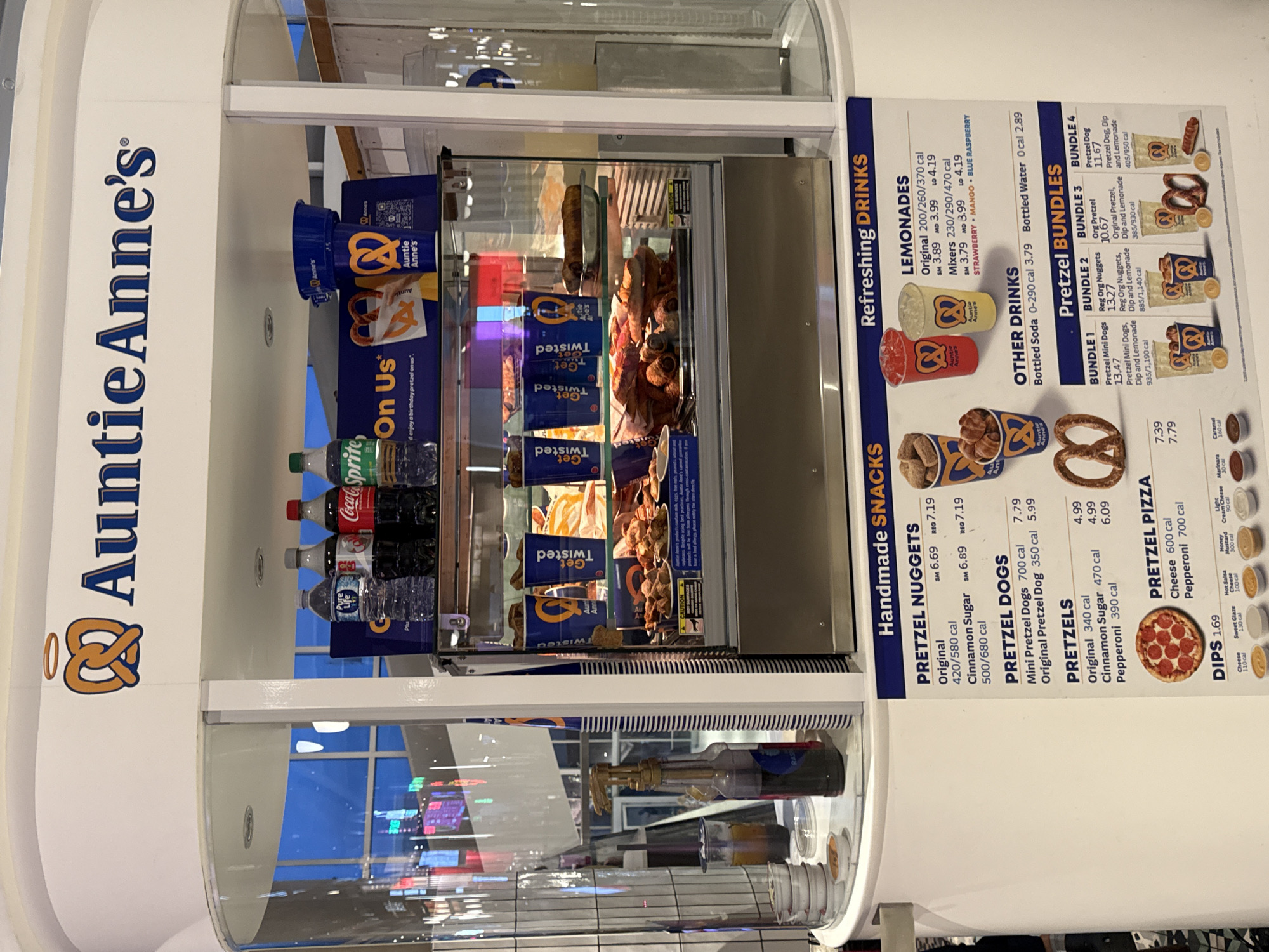 An Auntie Anne's pretzel stand is depicted in an indoor setting, possibly a mall or food court, evidenced by the reflective surfaces and artificial lighting. No people are visible.
The stand features a prominent "Auntie Anne's" logo and a display case showcasing various fresh pretzels, pretzel nuggets, and pretzel dogs. Below the main display, a refrigerated section holds bottled beverages like Coca-Cola, Sprite, and Pure Life water. Several branded cups and pretzel bags with "Get Twisted" text are also visible.
A detailed menu board on the right lists "Refreshing DRINKS" including lemonades (Original, Strawberry, Mango, Blue Raspberry mixers) and "OTHER DRINKS" (bottled soda and water). It also features "Handmade SNACKS" such as Pretzel Nuggets (Original, Cinnamon Sugar), Pretzel Dogs (Mini, Original), Pretzels (Original, Cinnamon Sugar, Pepperoni), and Pretzel Pizza (Cheese, Pepperoni). "DIPS" are also listed, including Hot Salsa, Sweet Glaze, Cheese, Honey Mustard, Light Cream Cheese, Marinara, and Caramel, all priced at 1.69.
Additionally, "Pretzel BUNDLES" are advertised, combining items like Mini Pretzel Dogs, Regular Original Nuggets, Original Pretzel, and Pretzel Dogs with dips and lemonades. Calorie information is provided for most menu items. The stand appears ready for business, with a condiment dispenser also visible on the lower left.