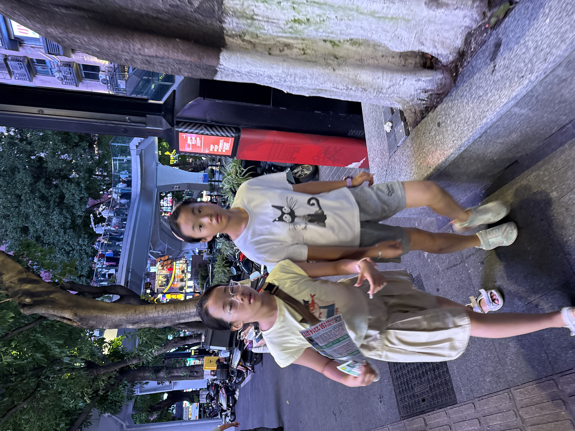 Two young girls are walking on a sidewalk at night. The girl on the left is wearing a light yellow t-shirt, a beige pleated skirt, and white sandals. She has glasses on and is holding a folded newspaper with advertisements. She is looking down at the newspaper and appears to be talking. The girl on the right is wearing a white t-shirt with a black cat graphic, grey shorts, and light blue sneakers. She is looking to her right and her mouth is slightly open. They are walking past a tree trunk with white and grey paint on it. In the background, there is a pedestrian overpass with people on it, buildings with lit windows, and parked motorcycles. The scene appears to be in a busy urban area, possibly near a commercial district, within the Yuzhong District of China.