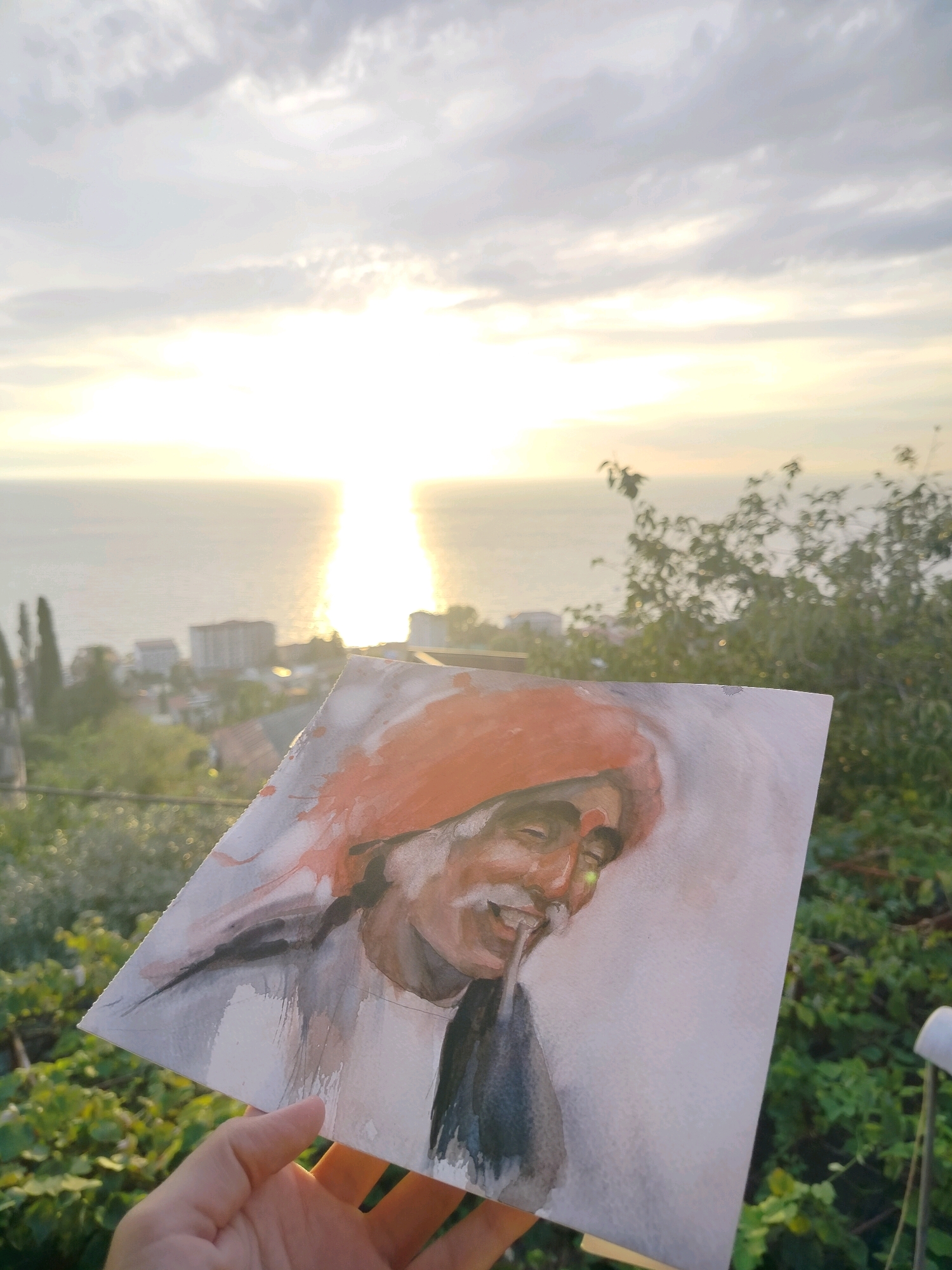 A watercolor painting of a man with an orange headdress is held up in front of a scenic sunset over the Black Sea. The painting is in the foreground, with a person's hand holding it from the bottom left. The man in the painting has a mustache and is smiling. The background shows a hazy sunset with bright sunlight reflecting on the water. The sky is filled with soft clouds. In the middle ground, there is a coastal town with buildings nestled amongst lush green trees and cypress trees. The scene appears to be a landscape from Gagra, Abkhazia. The time of day is sunset, and the weather appears clear with a gentle glow. The overall mood is peaceful and artistic, with the painting adding a personal touch to the natural beauty of the location.
