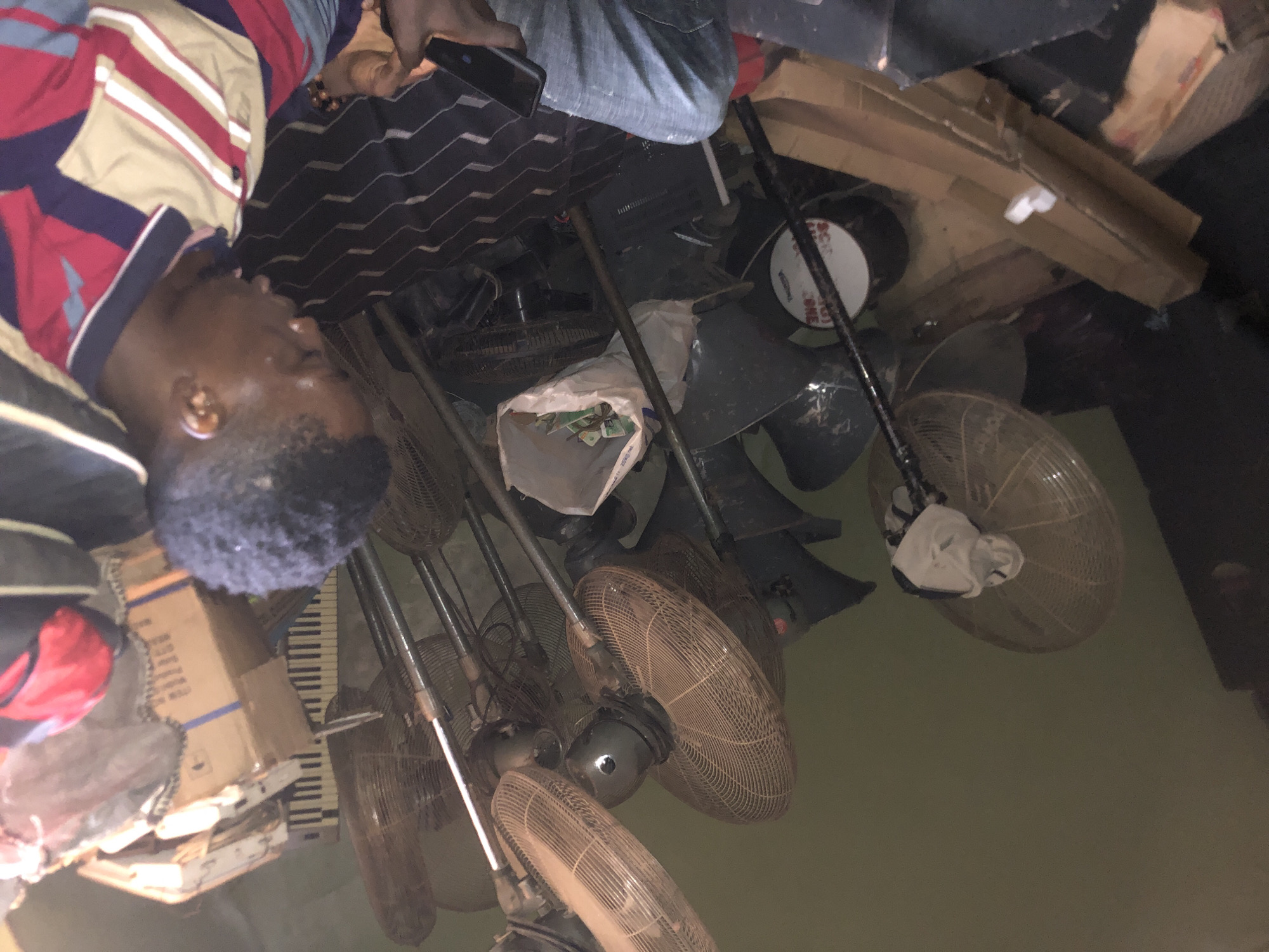 A man lies on the floor amidst a cluttered room in Nteje, Nigeria. The room is filled with numerous old fans, boxes, and other miscellaneous items. The man is wearing a striped shirt. He appears unconscious.