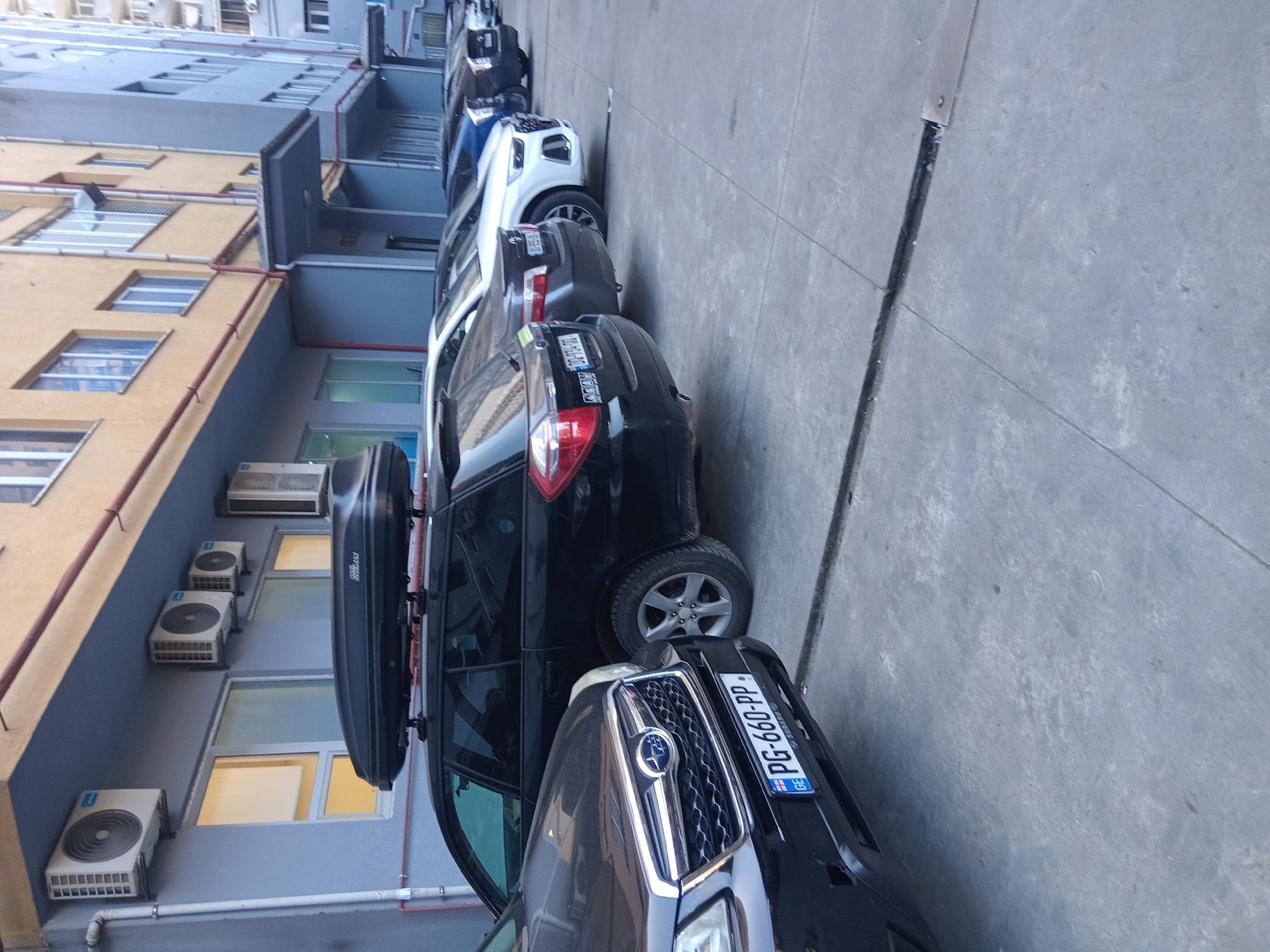 The image depicts a street scene with a row of parked cars alongside a multi-story building. The building has a yellow facade with grey balconies and numerous air conditioning units attached to its side. The parked cars, including a dark gray SUV with a roof cargo carrier, are lined up along a concrete sidewalk. The SUV's license plate reads "PG-660-PP" and indicates it is registered in Georgia. The image's perspective suggests it was taken at street level, possibly by someone standing on the sidewalk. The lack of people and other indicators makes it difficult to determine the precise location within Batumi, Georgia, beyond that it is likely a residential area with on-street parking.