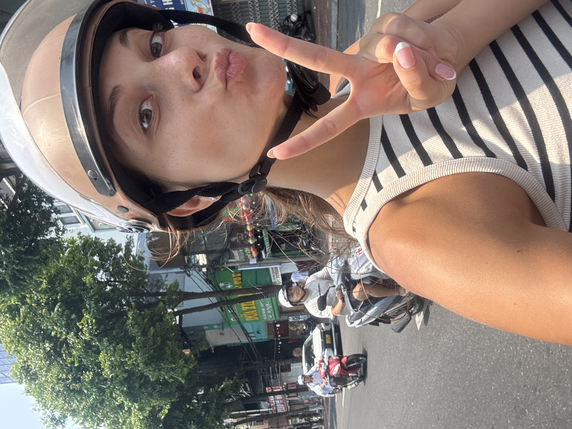 A woman wearing a brown helmet and a striped tank top is on a motorbike. She is holding up a peace sign. She is in Đà Nẵng, Vietnam. Other motorbikes and cars are visible on the road behind her. Buildings and trees are in the background.