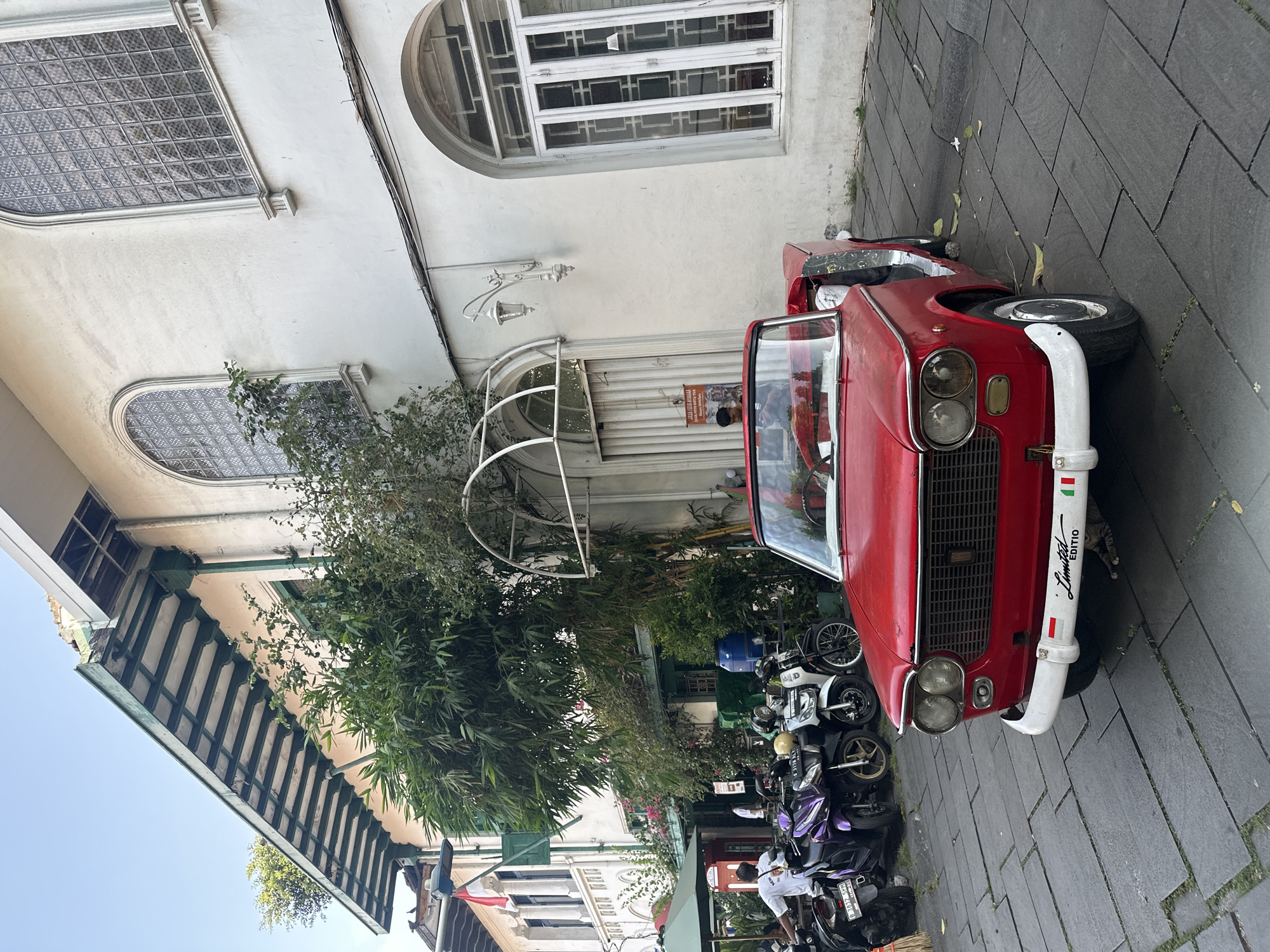 A red Fiat 124 Spider is parked on a gray-paved street next to a building with a white facade and arched windows. Numerous motorbikes are parked behind the car. A person wearing a white shirt stands beside one of the motorbikes. The photograph was taken in the city of West Jakarta, Indonesia.