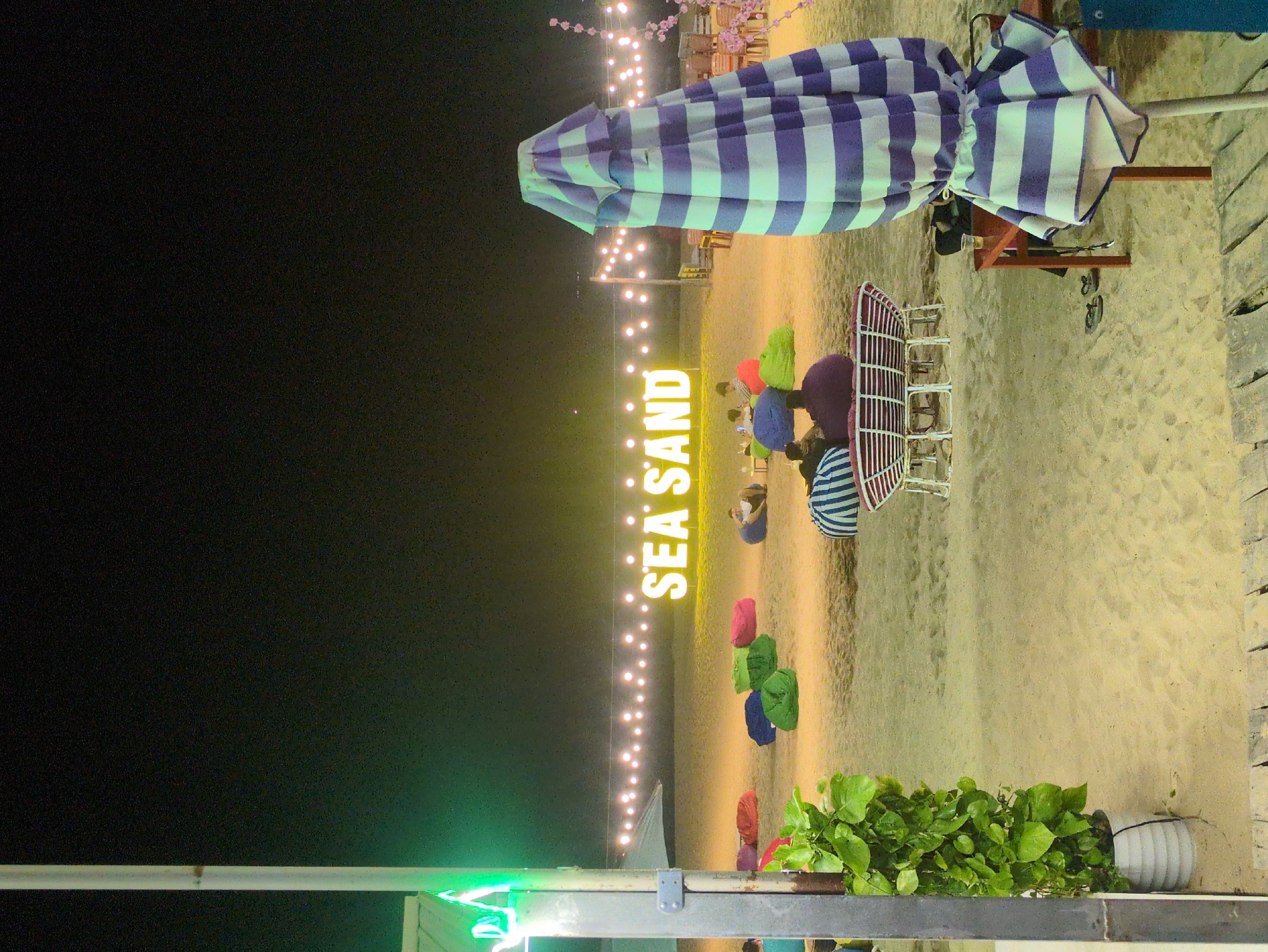 Captured at night in Quy Nhon Ward, Vietnam, the image shows a beach scene illuminated by string lights. A large "SEA SAND" sign glows prominently. Various bean bag chairs are scattered on the sand. There are people sitting on the beach. A striped umbrella is visible near a set of chairs. Green foliage and metal posts are in the foreground. The rest of the scene fades into darkness.