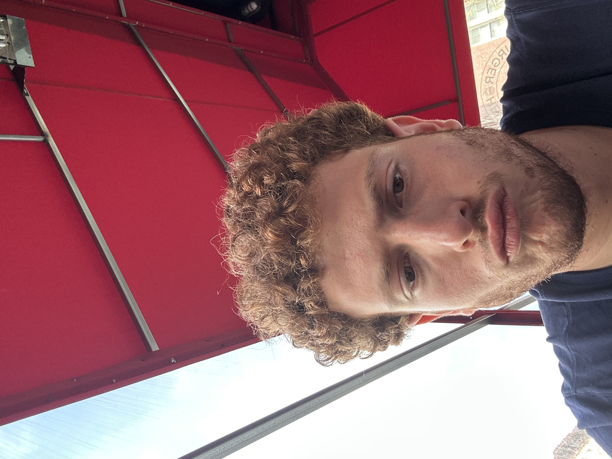 A medium close-up, portrait-oriented image, currently rotated 90 degrees clockwise, features a young man against a background of a vibrant red awning and a bright sky. The man, positioned in the center of the frame (when correctly oriented), has light brown, curly hair and a light beard or stubble. He is looking directly into the camera with a neutral expression, his lips slightly parted. He wears a dark blue top, visible on the right side of the frame.
The background is dominated by a large, red awning or canopy, supported by dark grey or black metal struts, which occupies the top and left side of the image. The fabric of the awning appears textured. Below and to the right of the man, a bright, clear, light blue sky is visible, suggesting daytime.
In the upper right corner, slightly obscured by the man's shoulder and the awning structure, there is a reflection or mirrored text visible on a distant window or building. The text is largely unreadable due to its mirrored and partial nature, but distinct characters such as "ЯКВСЯВЭН" or similar reversed letters can be partially discerned, hinting at a commercial establishment or sign. The overall setting appears to be an urban outdoor environment, possibly a street-side cafe or storefront in New York, USA, under an awning on a clear day.