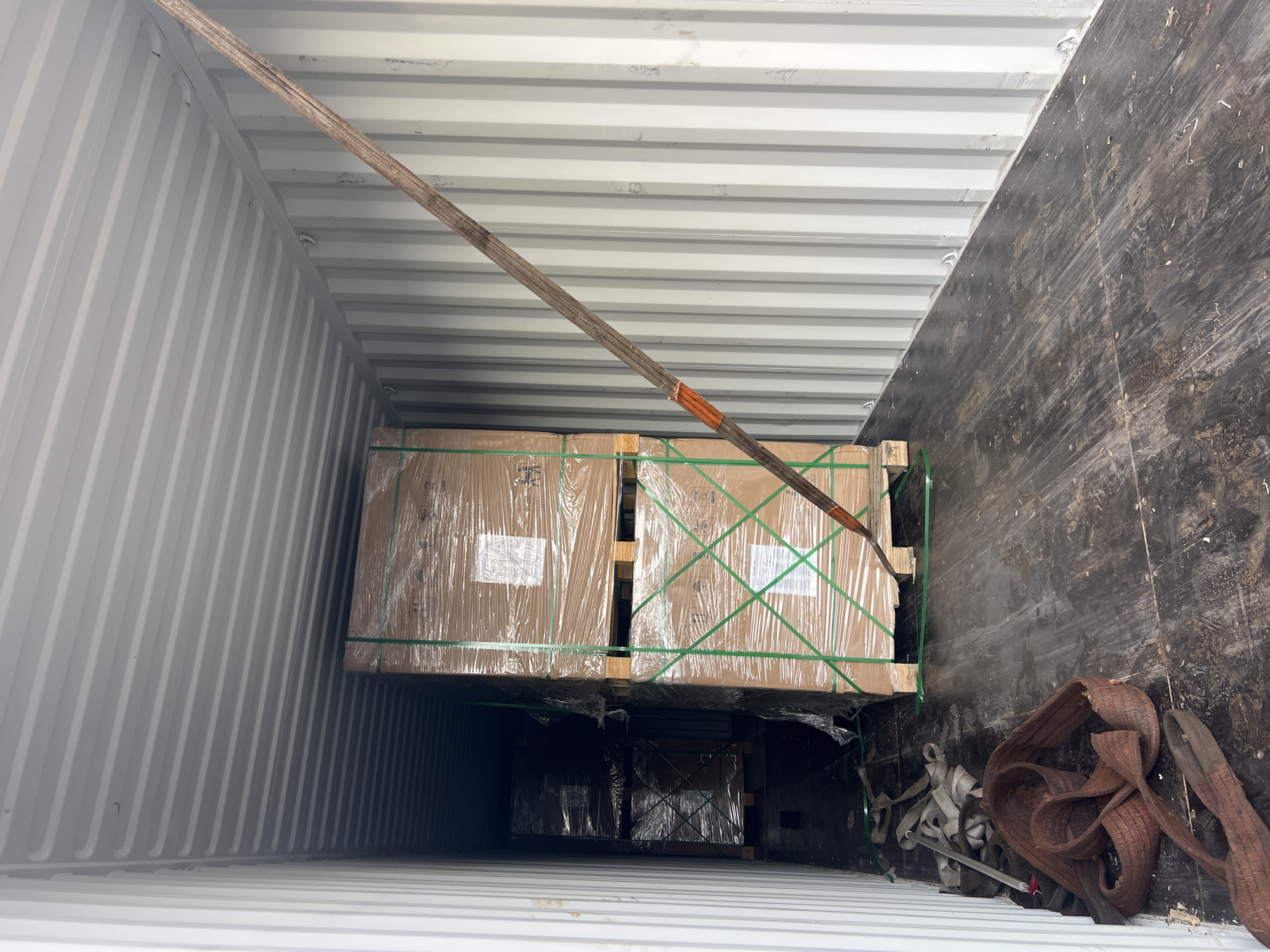 The interior of a shipping container, likely in a logistics or storage area, shows stacked pallets of goods secured with green strapping and wrapped in clear plastic. A thick brown strap runs diagonally across the top pallets, possibly used for securing or moving the load. Additional straps are piled on the right side of the container. The walls of the container are corrugated metal; the floor appears to be dark wood. The location is unknown.
