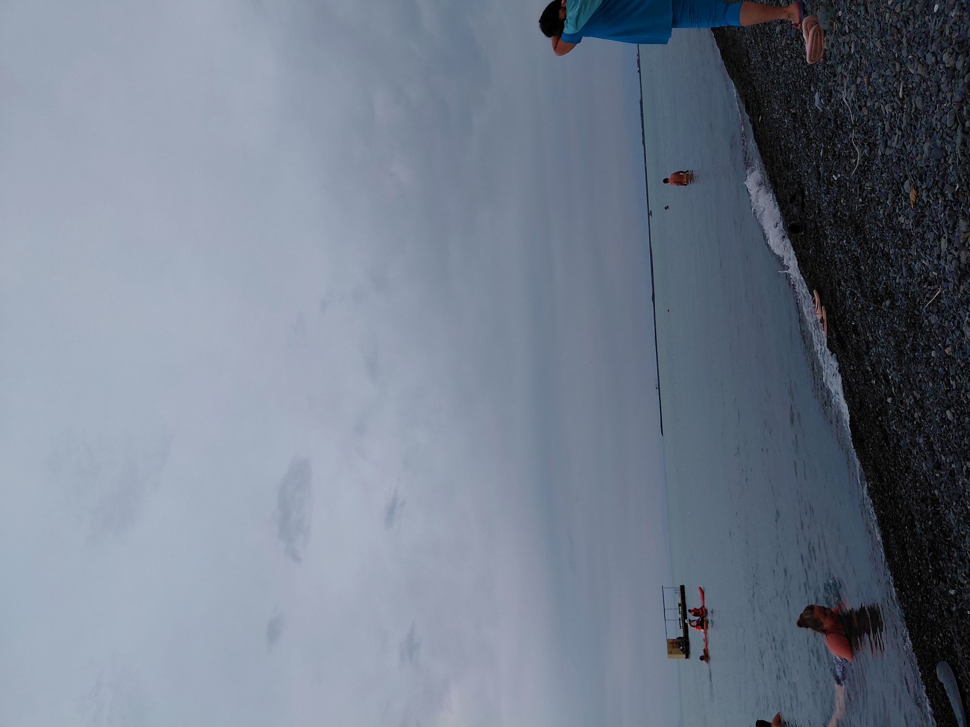 The image depicts a cloudy, overcast day at a pebble beach in Ақши, Kazakhstan. In the foreground, a person in a blue t-shirt and blue leggings stands with their back to the camera, their arms raised to their head. Scattered footwear, including sandals and shoes, lies on the pebble shore. To the left, a woman with her hair in a bun is partially submerged in the water, appearing to be swimming or wading. Further out in the water, a person stands in shallow water, and another is seen from the back in the water. In the mid-ground, a raft-like structure with a canopy is visible, with at least two individuals on it. Two orange kayaks are also present in the water near the raft, with at least two people in them, one appearing to be wearing a life vest. The sea stretches out to the horizon under a dense, gray sky. In the far distance, a thin, dark line suggests a distant shore or structure. The overall scene conveys a subdued, possibly cool, atmosphere with limited activity.