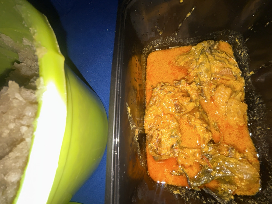 The scene depicts two distinct food items presented on a blue surface in Ilishan, Nigeria. In the upper portion, a light green bowl, partially cropped, contains a quantity of light-colored, granular fufu (likely pounded yam). Below it, a black rectangular plastic takeaway container holds a reddish-orange stew with several pieces of protein, possibly meat or fish, submerged within the sauce. The stew exhibits an oily sheen and a textured consistency. No individuals are visible. The items are arranged consistent with a meal ready for consumption.