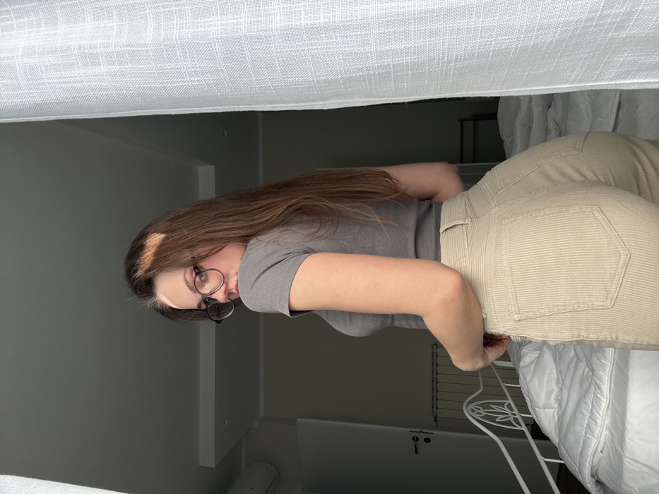 A female individual is depicted standing indoors in the city of Scafati, Italy. She is positioned with her back partially to the camera, her head turned over her left shoulder, looking directly forward. She has long brown hair and wears round eyeglasses, a grey ribbed short-sleeved top, and high-waisted beige corduroy pants. Her right hand rests near her lower back. The background features a white metal-framed bed with a white duvet to the left, a white radiator, a closed white door with a silver handle, and a wall-mounted air conditioning unit. A white translucent curtain occupies the right side of the frame, suggesting a window or light source. Natural light illuminates the scene from the right.