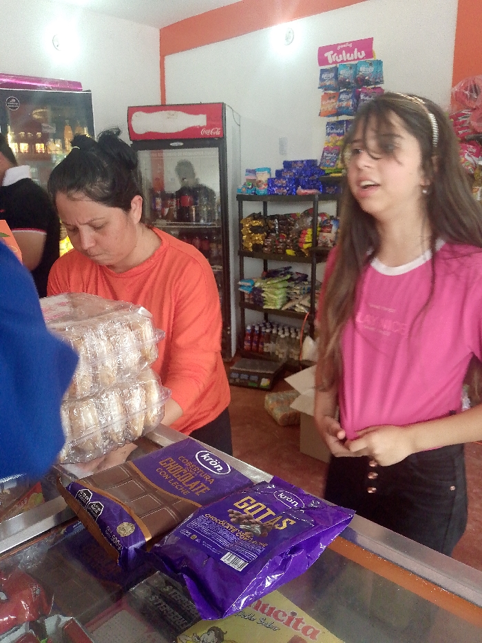 This indoor photograph captures a scene within a small store or bodega in Sabana de Machango, Venezuela. In the foreground, an older woman with dark hair tied up, wearing an orange long-sleeved shirt, is leaning over a counter. She is focused on handling two stacked clear plastic containers filled with what appear to be baked goods or pastries. Part of a person wearing a blue top is visible on the far left, suggesting they might be interacting with the older woman.
To the right, a younger woman with long, dark hair and a white headband stands upright, wearing a pink t-shirt and dark bottoms. She is looking to her right, mouth slightly open as if speaking or singing, with one hand resting near her waist. Her expression is animated.
The shop features a white wall with an orange trim along the top. In the background, a red Coca-Cola branded refrigerator is visible, reflecting the blurred image of a person inside. Beside the fridge, black shelving units are stocked with various packaged snacks, sweets, and bottles of beverages. Above one shelf, a display board with "Trululu" branding and assorted small bags of candy is mounted.
On the counter in the lower foreground, two purple packages of "Kron" brand chocolate are prominently displayed. One reads "COBERTURA CHOCOLATE CON LECHE" (Milk Chocolate Coating) and the other "GOTAS CHOCOLATE" (Chocolate Drops). Other visible text includes "Coca-Cola" on the fridge, "500g" on packaging, and partial text like "LAY NICE," "STEB," "ucita," and "Doble Sabor" on items further down. The overall setting is a brightly lit, typical local convenience store.