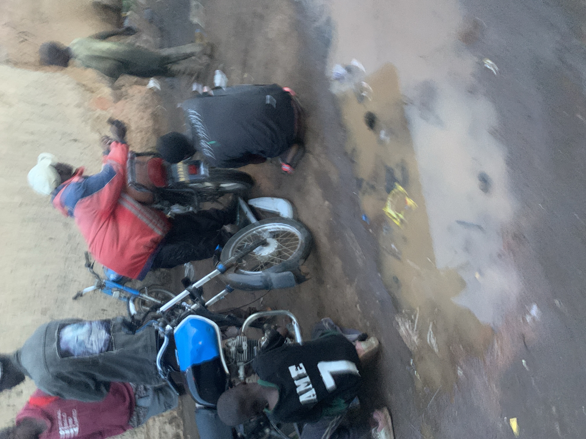 This image captures an outdoor scene in Jos, Nigeria, featuring multiple individuals and motorcycles on an unpaved surface. The ground consists of dirt and sand, interspersed with muddy patches and puddles, suggesting recent rain or poor drainage.
At least five people are present. In the upper left, one individual lies prone on the sandy ground, wearing a light-colored long-sleeved shirt and dark trousers. Towards the middle-left, a person wearing a red jacket with blue sleeves and a light head covering is situated near a red and silver motorcycle. Another individual in a dark top and trousers is seen bent over in the upper-middle section, near a dark-colored motorcycle.
In the lower-left, a person wearing a dark grey t-shirt with a white graphic print and dark trousers is seated or crouching beside a blue bicycle and another motorcycle. Adjacent to them, another person, wearing a black t-shirt with the white text "AME" and a large number "7" (or letter "L") on the back, is bent over, seemingly attending to the engine of a motorcycle.
Various pieces of debris are scattered across the ground, particularly around the wet areas. The lighting is soft and diffused, indicating an overcast sky or an early/late time of day. The activity primarily involves people gathered around motorcycles, with one person appearing to work on a bike, while the individual lying on the ground adds an ambiguous element to the scene. No distinct emotions are discernible from the individuals.
