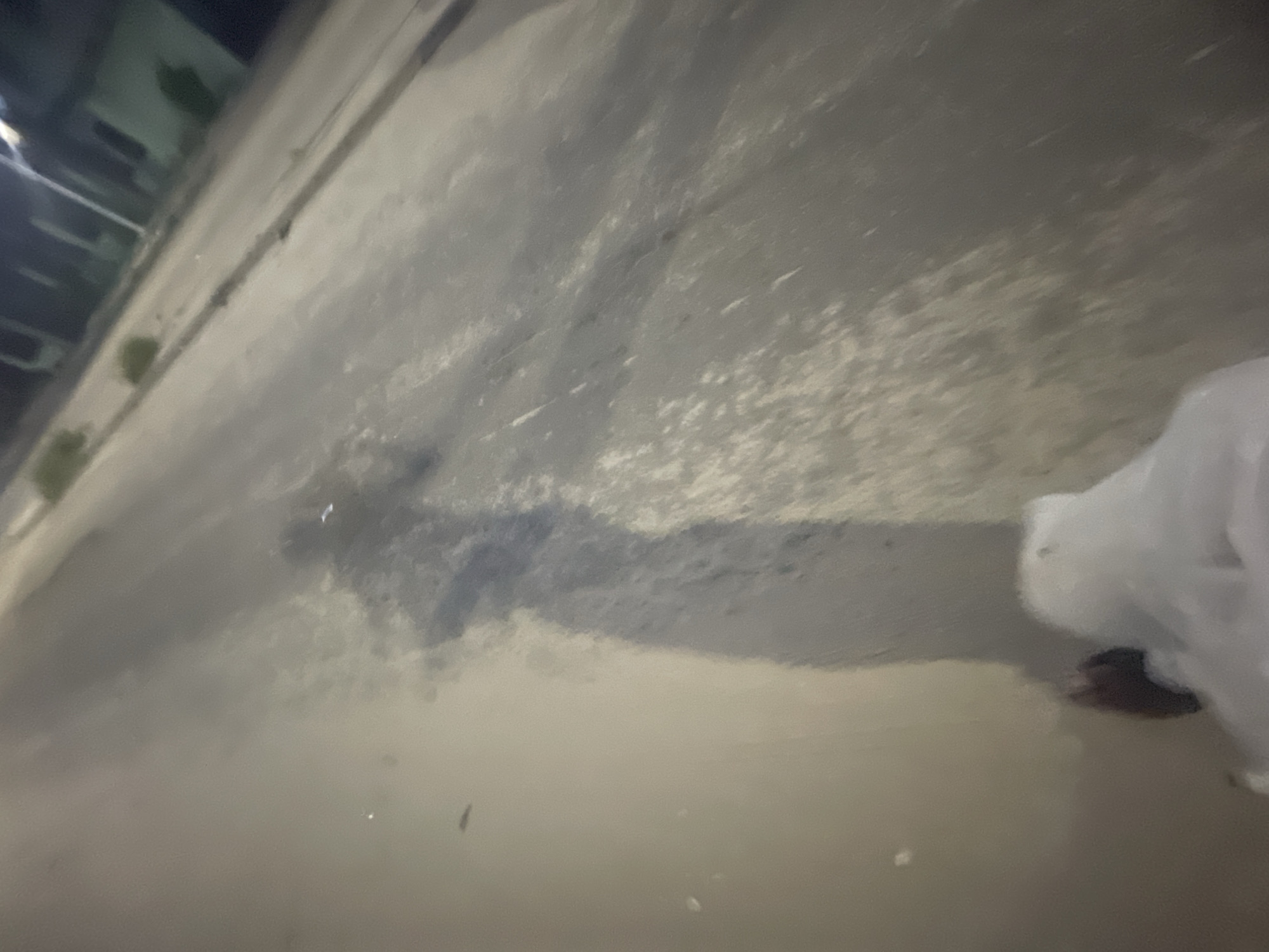 An image of a street in what could be Ilado, Nigeria, based on the prompt, but unconfirmed by visual information. The photo is taken at a low angle, pointed downwards onto a paved road with patchy dark streaks. In the upper left, a blurry building structure is visible with some green vegetation. A translucent white plastic bag is in the lower right corner, partially obscuring a person's foot wearing a brown sandal.
