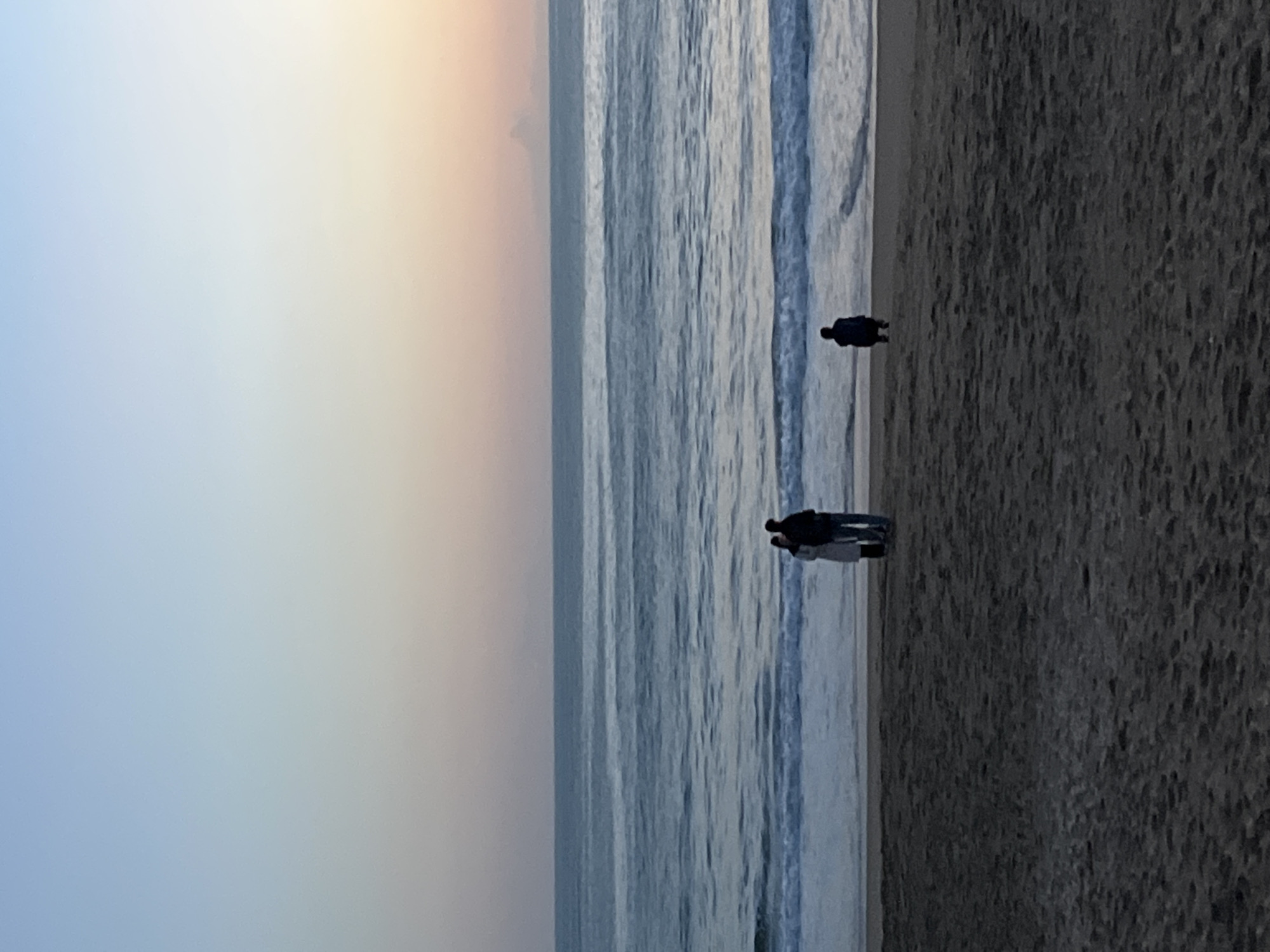 The image shows a beach scene, possibly in Alanya, Turkey, given its coastal location. The sky has a gradient of blue to peach, indicating either sunrise or sunset. The sea has gentle waves. There are two groups of people on the beach. One individual is standing closer to the water's edge, and a small group of three or four individuals is standing further down the beach. The sand appears dark.