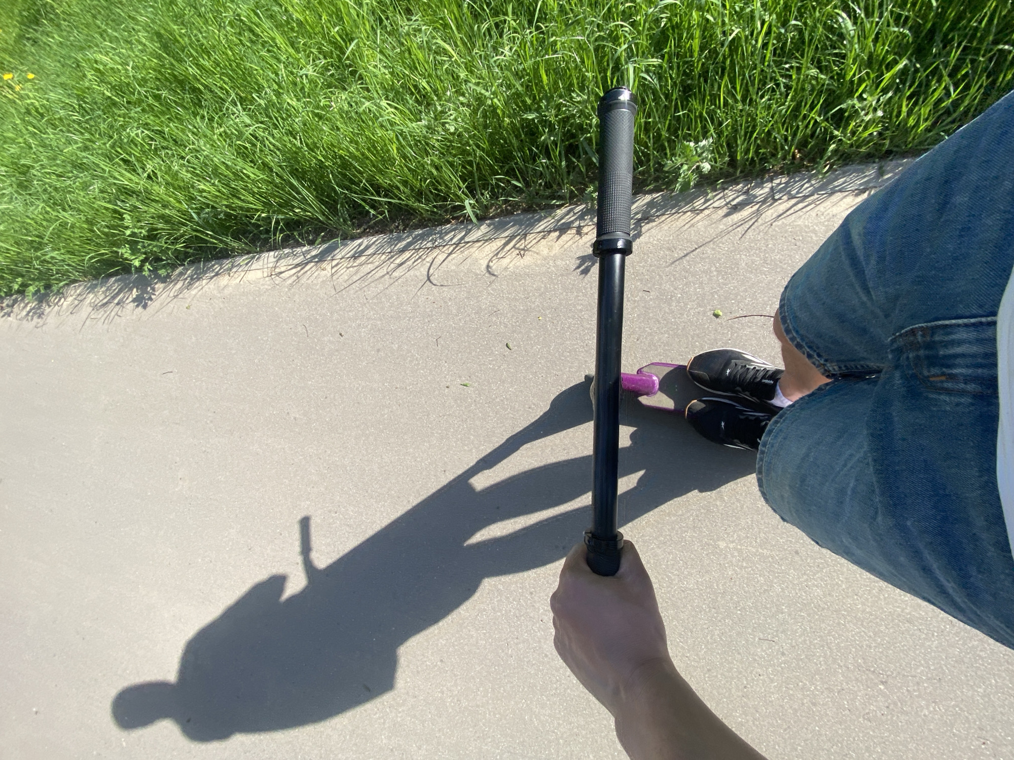 A first-person perspective image shows a person riding a scooter on a concrete path. The path is bordered by green grass on the left side of the frame. The person is wearing denim shorts and black athletic shoes with white socks. The scooter has black handlebars with textured grips and a pink deck. The shadow of the person and the scooter is visible on the concrete path. The location appears to be an outdoor public space, potentially a park or greenway.