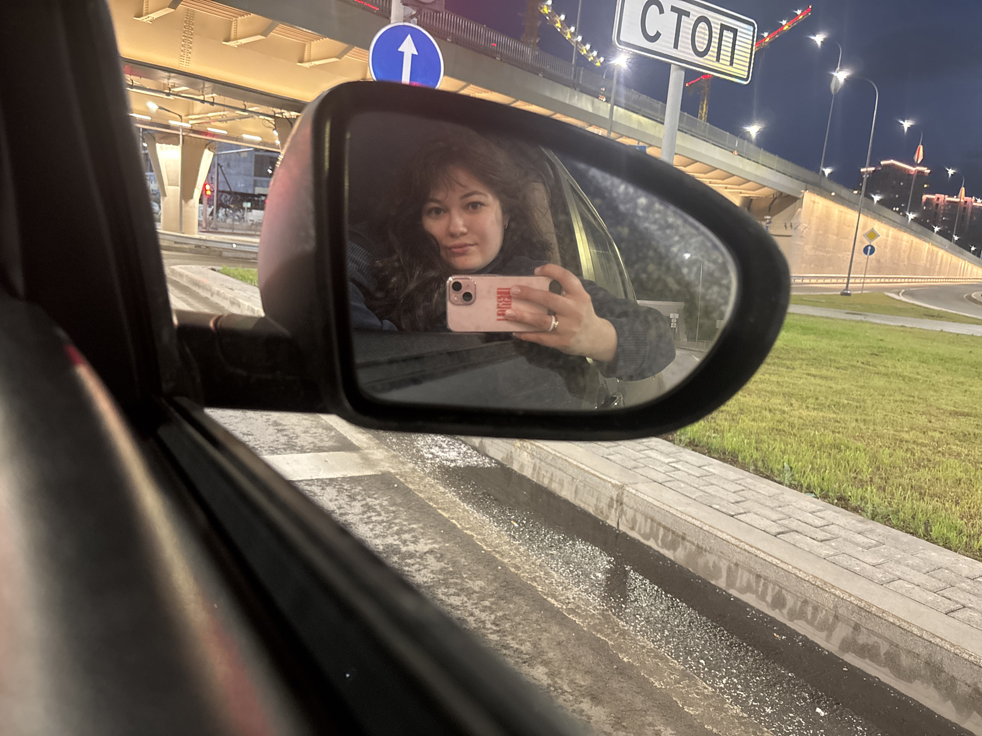 The image captures a nighttime urban scene from the interior of a car, with the primary subject being a woman taking a selfie reflected in the passenger-side mirror. In the foreground, the dark, blurred structure of the car's door frame and window runs along the left side and bottom of the image. The central element is the car's passenger-side mirror, which reflects a woman. She has long, dark, wavy hair and is wearing a dark, possibly knitted, garment. She holds a light pink iPhone, likely an iPhone 13 or 14 Pro based on its camera array, in her left hand, positioned to capture her reflection. A red sticker, which appears to say "REDLAB" in mirrored text, is visible on the phone case. A ring is worn on her left ring finger. Her gaze is directed towards the phone, capturing the selfie. Beyond the car and reflected in the mirror, the background reveals a significant road interchange under dusk or night conditions. A large concrete overpass or bridge dominates the upper and middle sections of the scene, illuminated from beneath with warm yellow-orange lighting that highlights its structural beams. Distinctive streetlights with a multi-pointed star-like glow are visible along the road and extending towards the distant city. A prominent concrete support pillar for the bridge stands below the overpass. A red traffic light can be seen under the bridge on the far left. Road signage is clearly visible: a rectangular white sign with the black Cyrillic text "СТОП" (STOP) is positioned high above the bridge, and a circular blue sign with a white arrow pointing upwards (indicating compulsory straight ahead) is located below it, closer to the viewer. In the far distance, across and beyond the bridge, modern high-rise buildings with illuminated windows and red or orange architectural accents are visible against the dark sky. A red construction crane is also discernible behind the "СТОП" sign, suggesting ongoing development. At ground level, a paved road surface is visible, alongside a concrete curb and a strip of green grass. The area immediately adjacent to the curb appears wet or covered in a thin layer of dirty water or slush, reflecting the artificial lights. This scene is located in **Perm, Russia**, specifically depicting the modern road interchange leading to the Chusovoy Bridge on the right bank of the Kama River, or a nearby major junction within the city, identifiable by the Cyrillic signage,