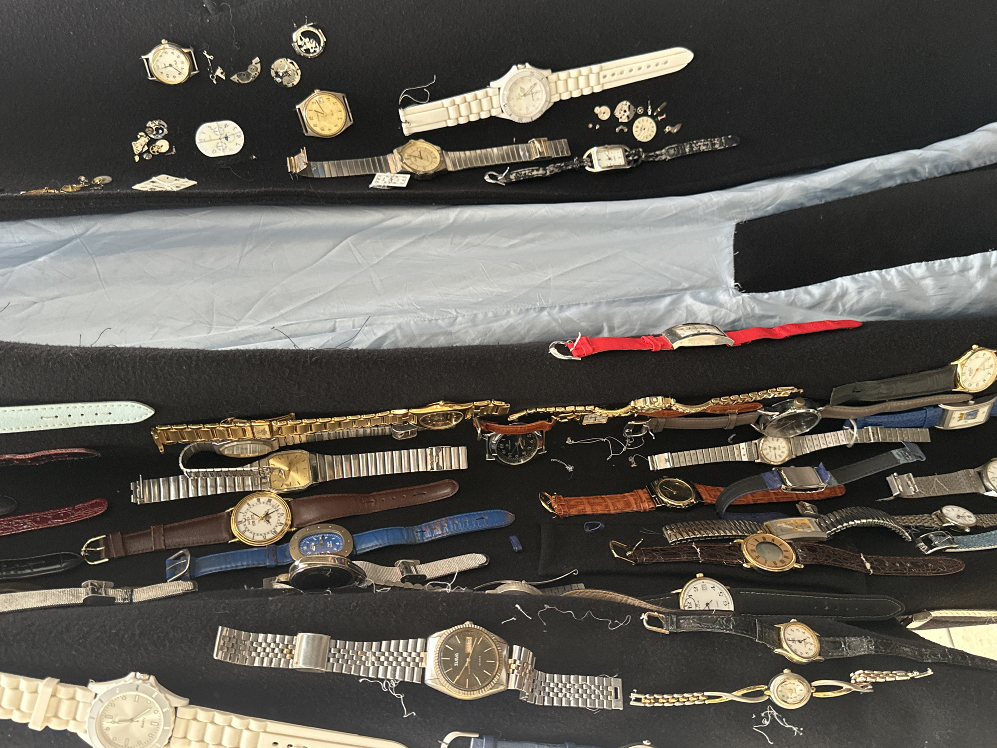 The image shows a collection of watches, watch parts, and watch straps arranged on a black fabric surface. Some are intact watches with bands, while others are disassembled, with parts scattered around. A blue-grey material is visible beneath the black fabric in the upper part of the image. The scene appears to be a disorganized collection of timekeeping devices and related accessories. There is no information available in the image to suggest a location in the city of Montreuil, France.