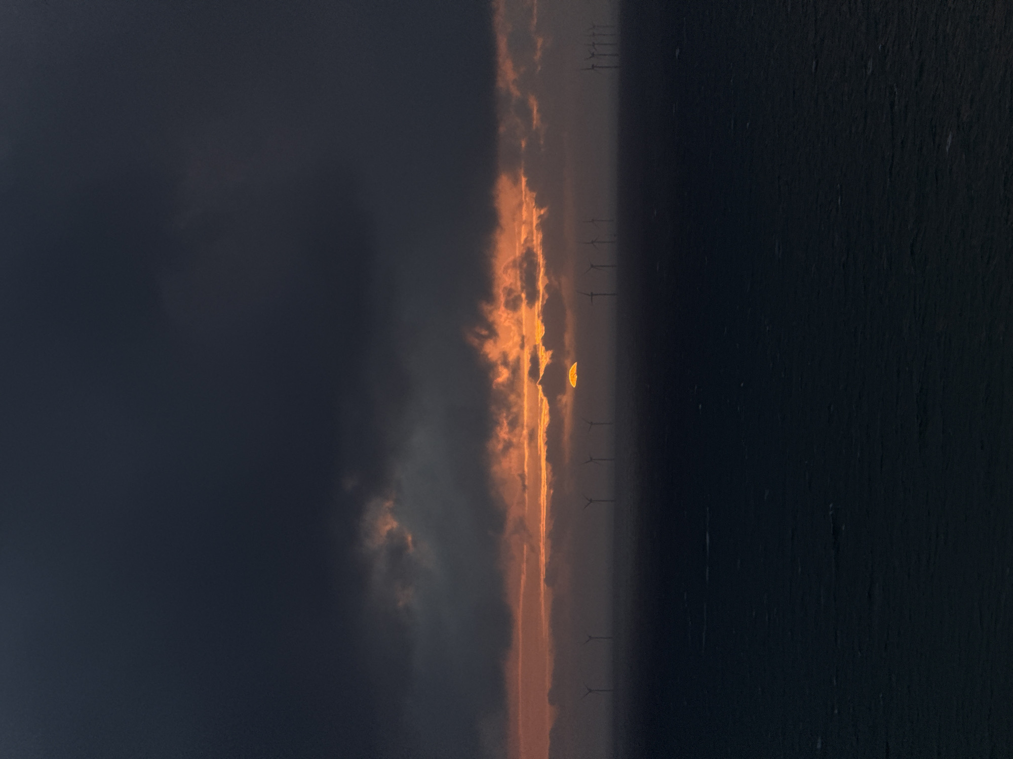 The image captures a dramatic sunrise or sunset over the ocean, showcasing an offshore wind farm. The sky is dominated by a sweeping band of vibrant orange and yellow clouds, horizontally bisecting the frame. This luminous strip, suggesting the sun's low position, is bordered by heavy, dark grey and deep blue clouds above, creating a stark contrast. Below the fiery horizon, the dark, calm surface of the ocean extends into the foreground.
Silhouetted against the brilliant light and stretching across the water are numerous wind turbines. Their distinct shapes, including the central tower and the blades, are clearly visible as dark forms against the glowing sky. The turbines appear to be arranged in a line, emphasizing the scale of the offshore wind farm.
The time of day is clearly either sunrise or sunset, indicated by the intense colors of the sky and the low angle of the light. The weather appears partly cloudy, with a clear horizon allowing the sun's rays to illuminate the cloud band. The scene conveys a sense of serene power, blending natural beauty with human engineering for renewable energy. The location is an offshore wind farm near Fécamp, France. No visible text is present in the image.
