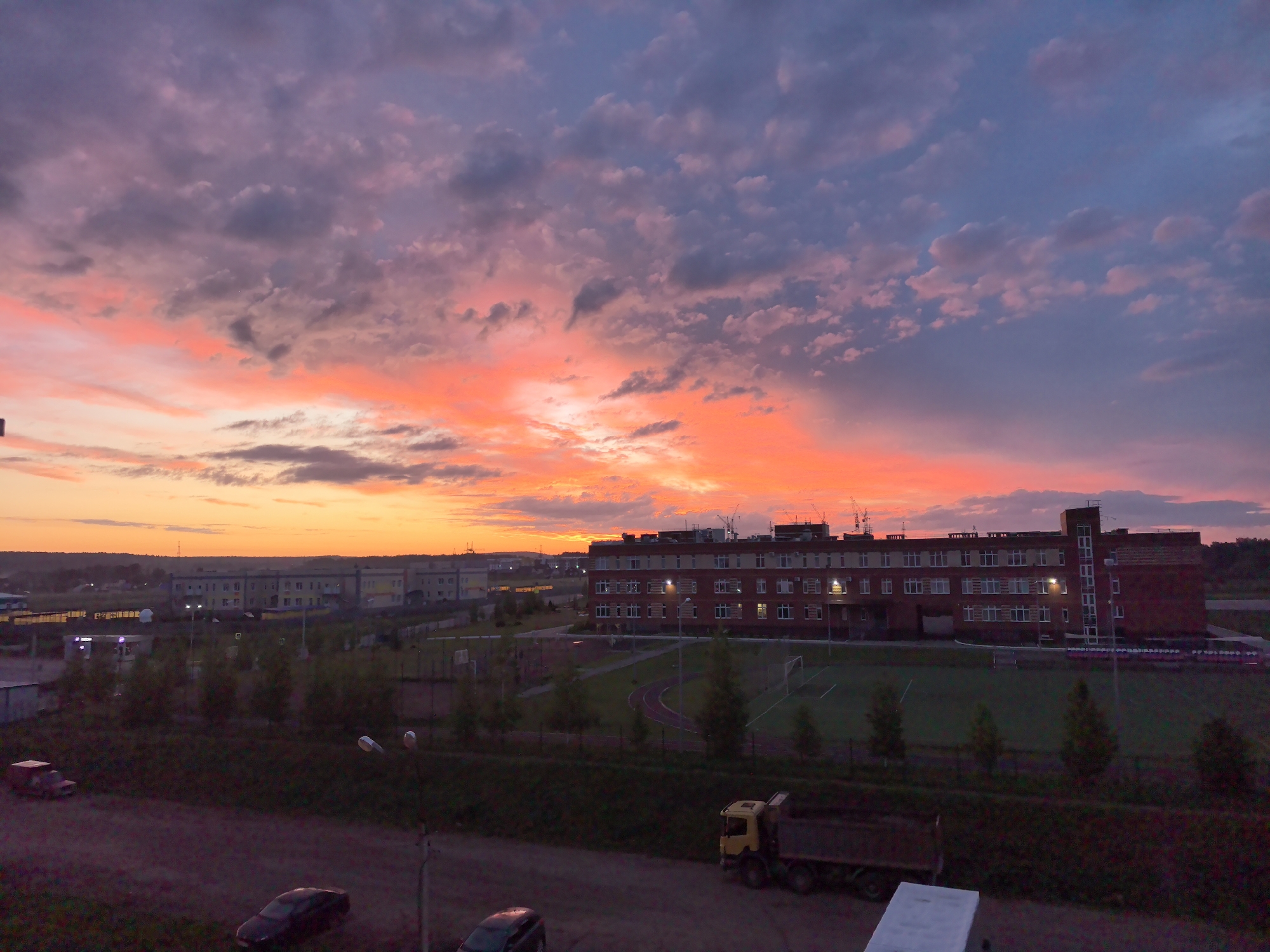 The image depicts a view of Фролы, Russia at dusk or dawn, characterized by a vibrant, multicolored sky transitioning from orange and yellow on the horizon to pink and purple overhead, with scattered clouds throughout.
A multi-story brick building with symmetrically placed windows is situated near a green space containing a running track and a soccer field. In the foreground, a parked yellow dump truck and several cars are visible on a dark, unpaved surface.
In the background, another building is observed along with additional signs of urban infrastructure.