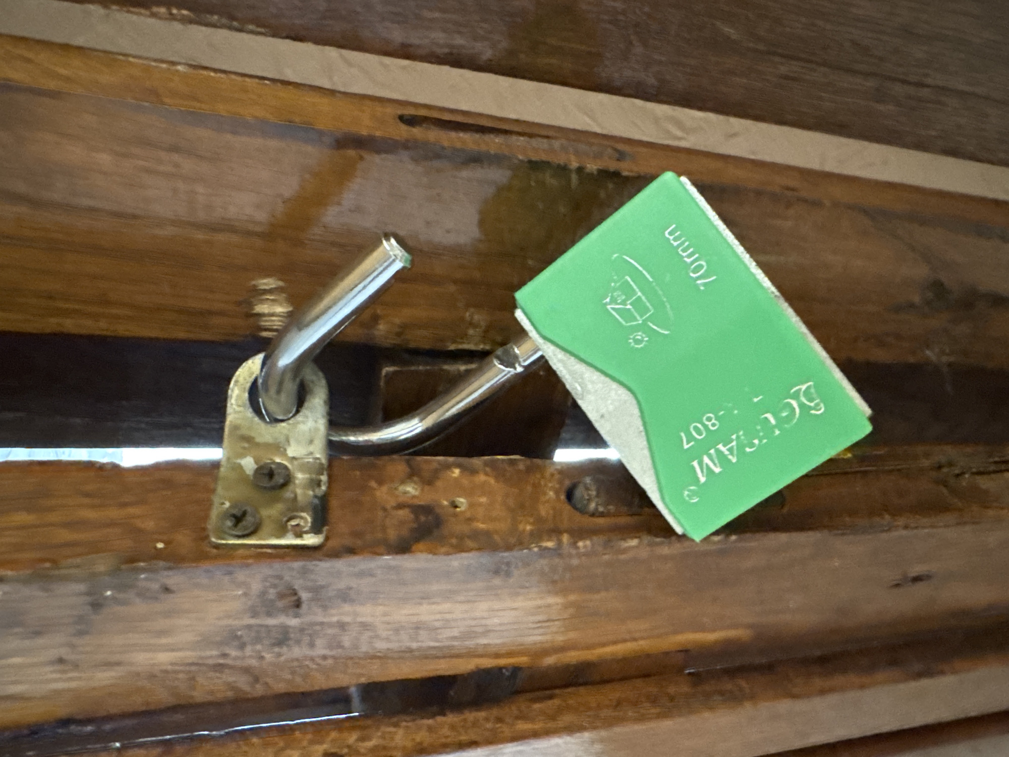 The image shows a close-up of a wooden surface, likely a chest or cabinet, with a metal latch mechanism. The latch is partially open, attached to the wood with two screws. A small, bright green object, possibly a product tag or label, is leaning against the latch. The green object has text printed on it, which is partially visible and appears to be in English. The visible text includes "70mm", "SEUGAM", and "-807". The wooden surface exhibits a visible grain pattern and appears to be aged. The lighting suggests an indoor setting, possibly during the day. The scene is static, with no people or active events depicted. The context provided indicates the location as Odesa, Ukraine.
