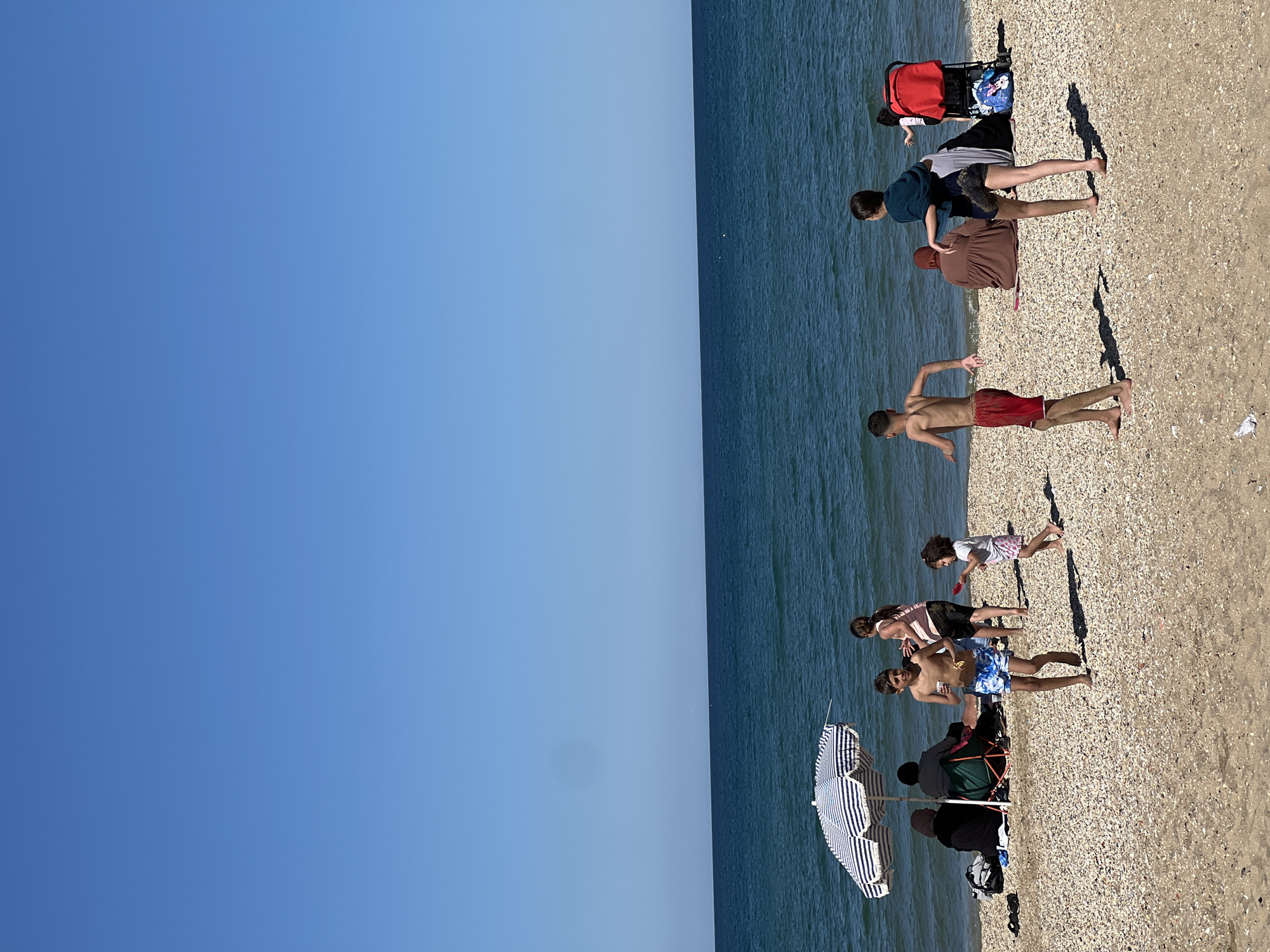 The image depicts a beach scene in M'Diq, Morocco, with a clear blue sky above a body of water meeting the sandy shoreline. Several people are present on the beach. At least one person is seen standing in the water's edge. Another is walking in the beach. There is a group with a striped beach umbrella. Some people appear to be relaxing near a beach chair. A few individuals are interacting or playing in the shallow water. The beach itself has a mix of sand and pebbles.