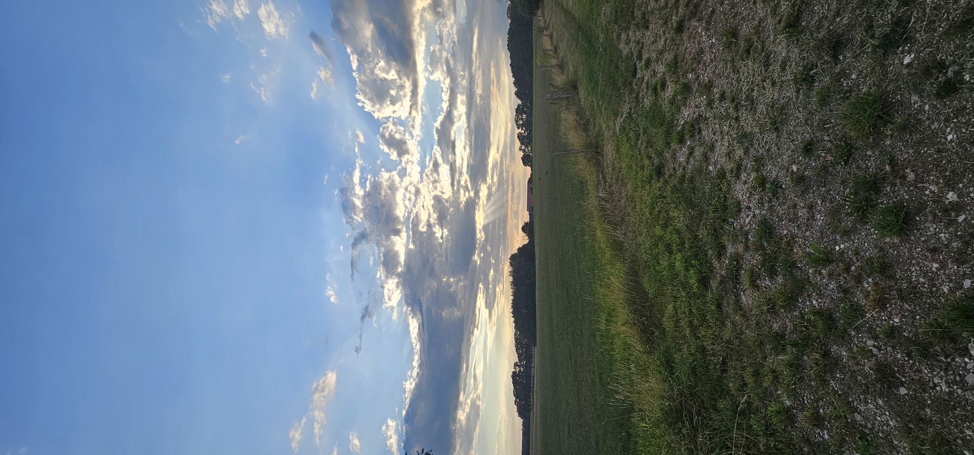 This is a vertical, wide-angle photograph of a rural landscape in Groenlo, Netherlands, during what appears to be late afternoon or early evening. The sky dominates the upper two-thirds of the frame, filled with a mix of blue sky and scattered clouds. Sunlight is breaking through the clouds in visible rays, illuminating portions of the sky and casting subtle shadows.
The horizon is low in the frame, demarcated by a line of trees and a distant building with a red roof on the left side. The land in the middle ground is a flat, open field of green grass, stretching from left to right. The grass is lush and varies in shade, with some taller, drier blades visible in the foreground on the left.
A rough, gravel path runs along the right edge of the frame, leading away from the viewer. Along the edge of the field and path, there are several wooden fence posts, some with barbed wire attached, indicating a boundary or enclosure. The ground along the path is a mix of dirt, small stones, and sparse vegetation. The overall impression is one of tranquility and natural beauty, with the setting sun creating a dramatic and serene atmosphere.