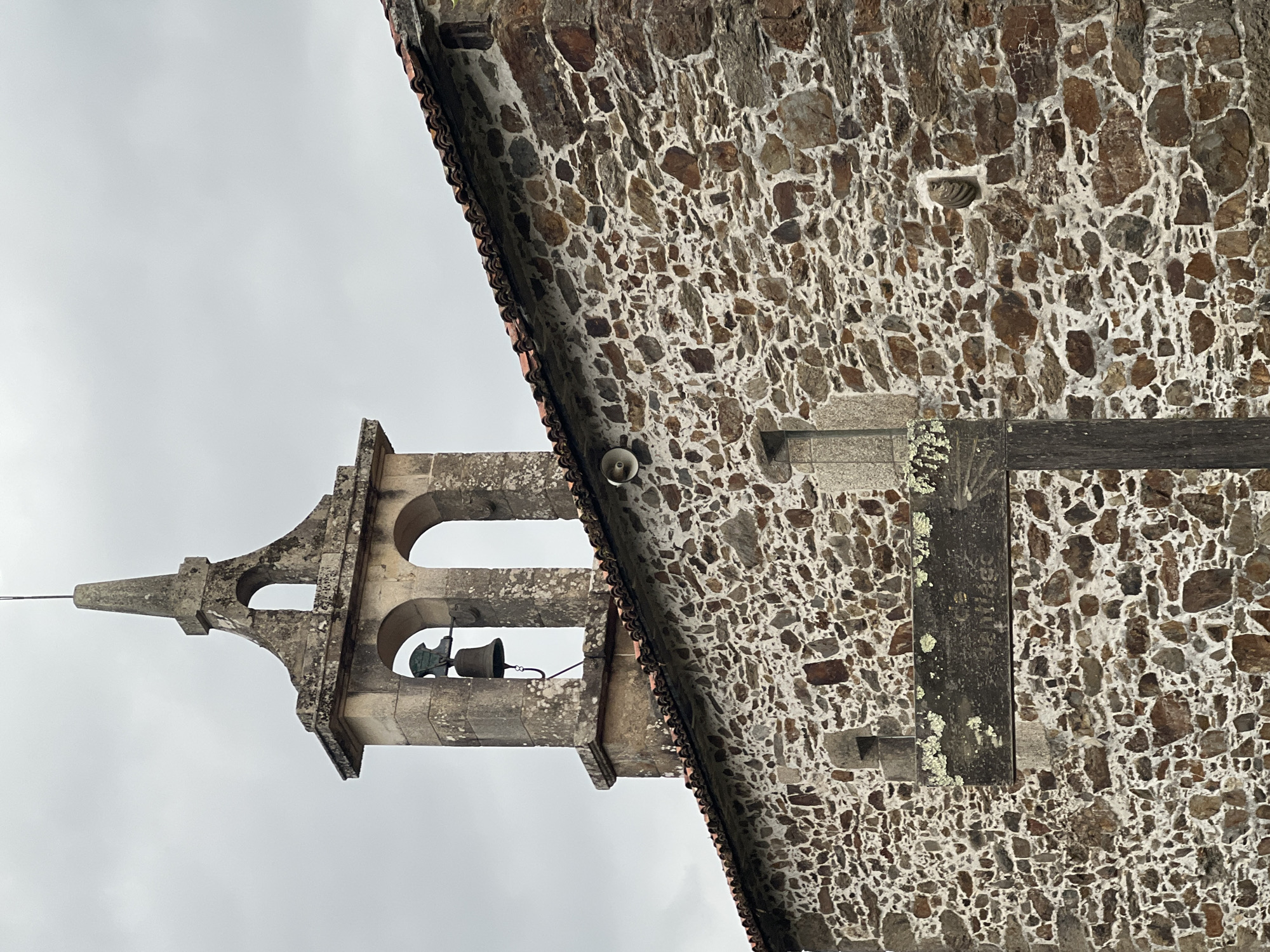 The image shows a stone church in San Paio, Spain, with a bell tower and a tiled roof. A wooden signpost with moss-covered lettering is mounted on the stone wall of the church. The signpost points to the right, and the visible text appears to say "Santiago." A bell hangs in the arched opening of the bell tower, which is topped with a pointed spire. The sky is overcast. The building is constructed of rough-hewn stones of various sizes and colors, mortared together with white cement. A small, circular speaker is mounted on the wall to the left of the signpost.