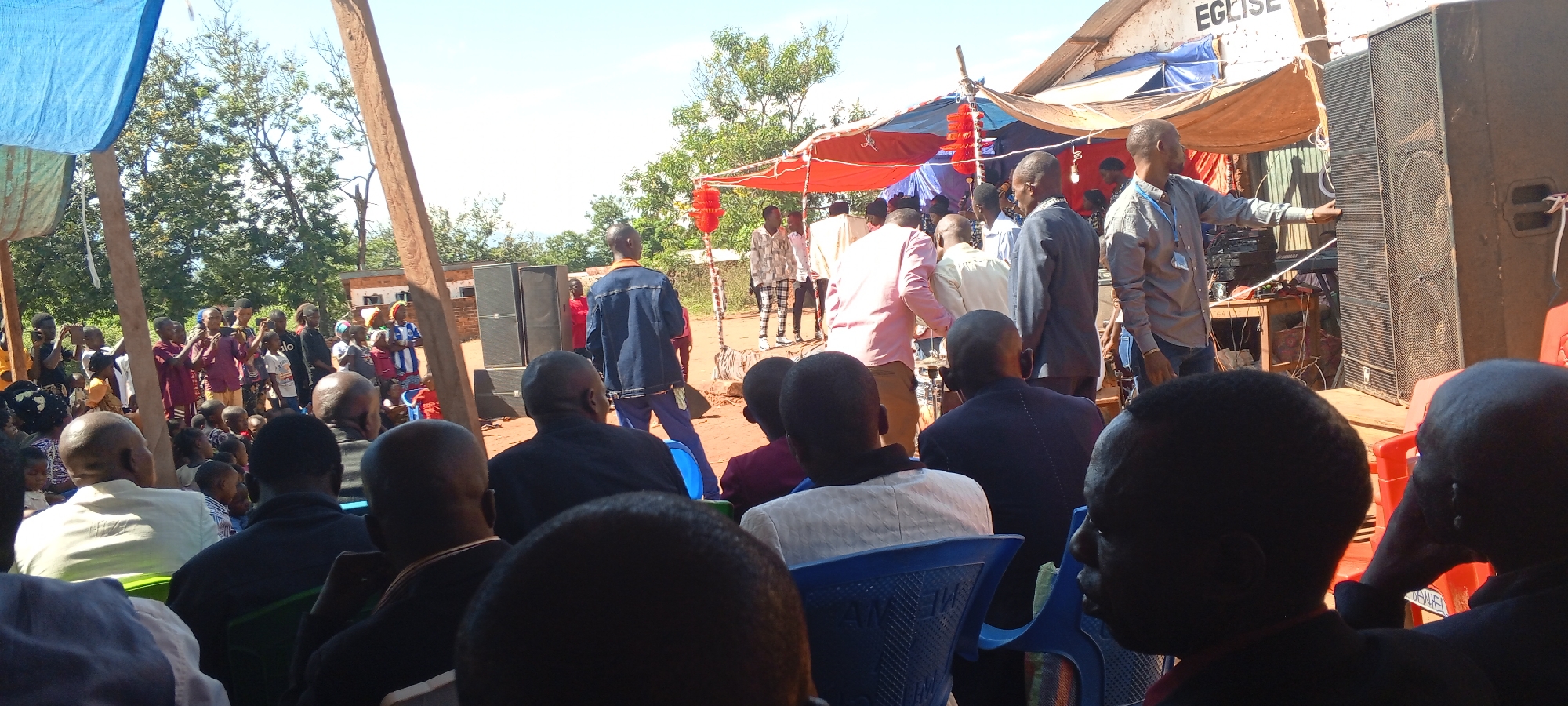 A daytime outdoor event in Kalemie, Democratic Republic of the Congo, features a crowd of people gathered near a building with "EGLISE" visible on its facade. Many individuals are seated facing a makeshift stage area covered with draped materials. Loudspeakers stand prominently on the stage, and a man in a gray shirt gestures towards one of them. The audience consists of a diverse group of people, including adults and children, some of whom are taking photos or videos with their phones. The environment includes trees and other buildings in the background, suggesting a community gathering.