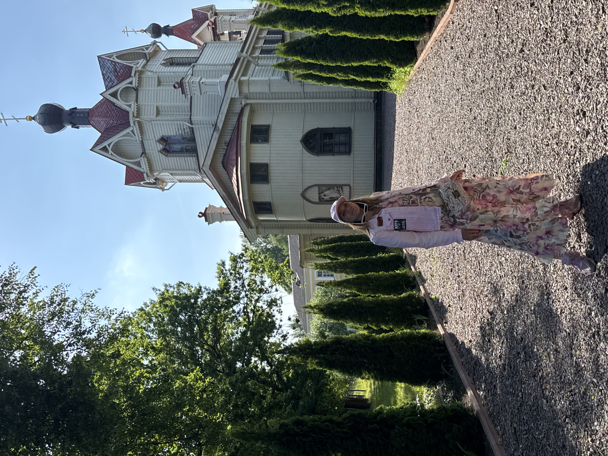 A woman stands on a gravel path in front of a wooden Orthodox church in Lomonosov, Russia. The church is white with red trim and features multiple domes topped with crosses. The woman is wearing a long floral dress, a light pink jacket, and a pink cap. She is accessorized with sunglasses and a small crossbody bag. The path is lined with manicured evergreen trees. The environment appears to be a sunny day with a clear blue sky.