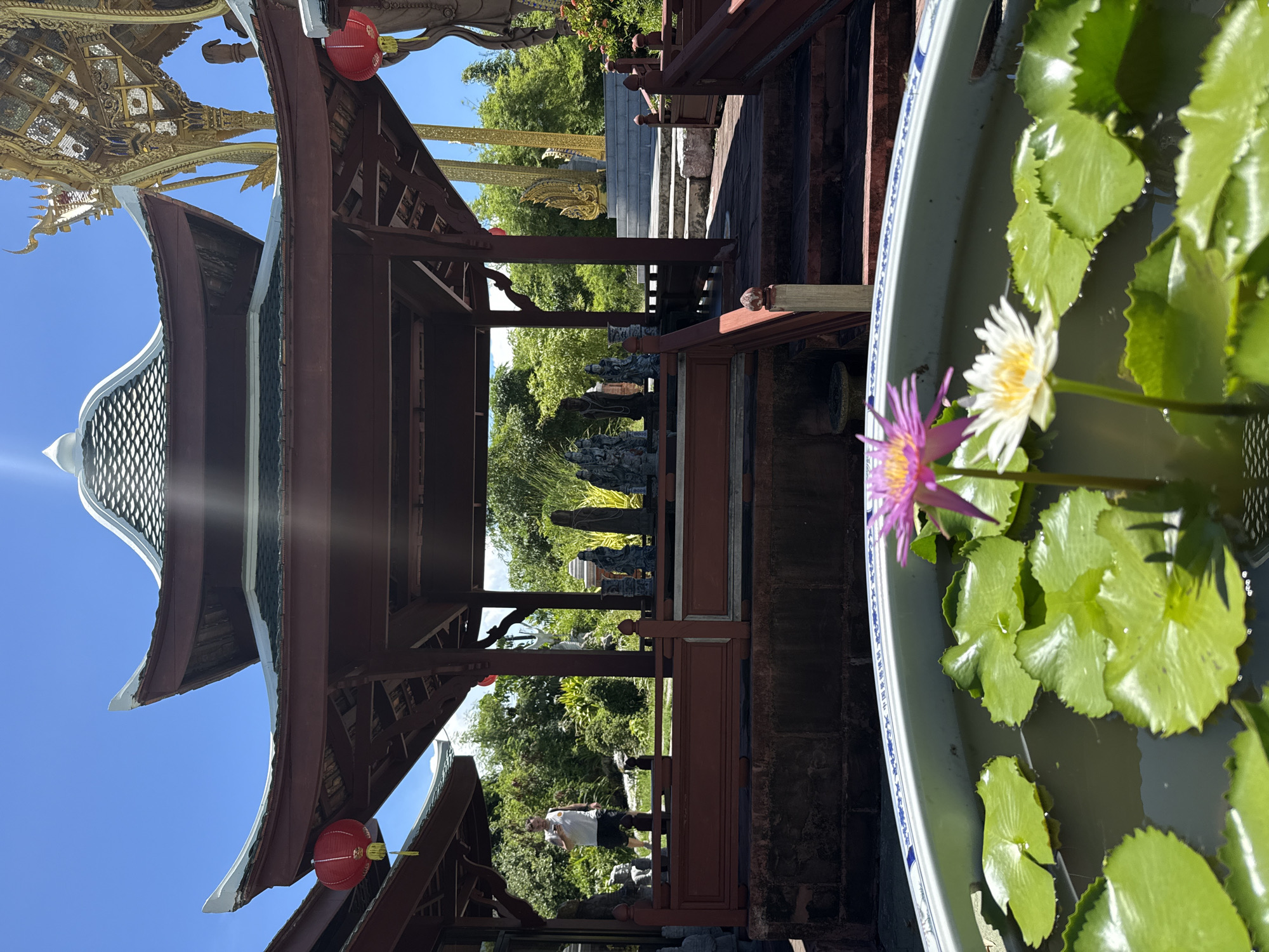 A vertical, eye-level shot reveals a pagoda at Ancient City in Bang Pu Subdistrict Municipality, Thailand. The shot is composed of a wooden pagoda on the left, a lotus-filled container on the right, and a garden in the background. The pagoda has a multi-tiered roof and red lanterns. The container is filled with water and lotus leaves, and two lotus flowers, one white and one purple, are in bloom. The garden is green and lush, with various statues and trees. A man can be seen walking within the pagoda's structure. The sky is blue and cloudless.